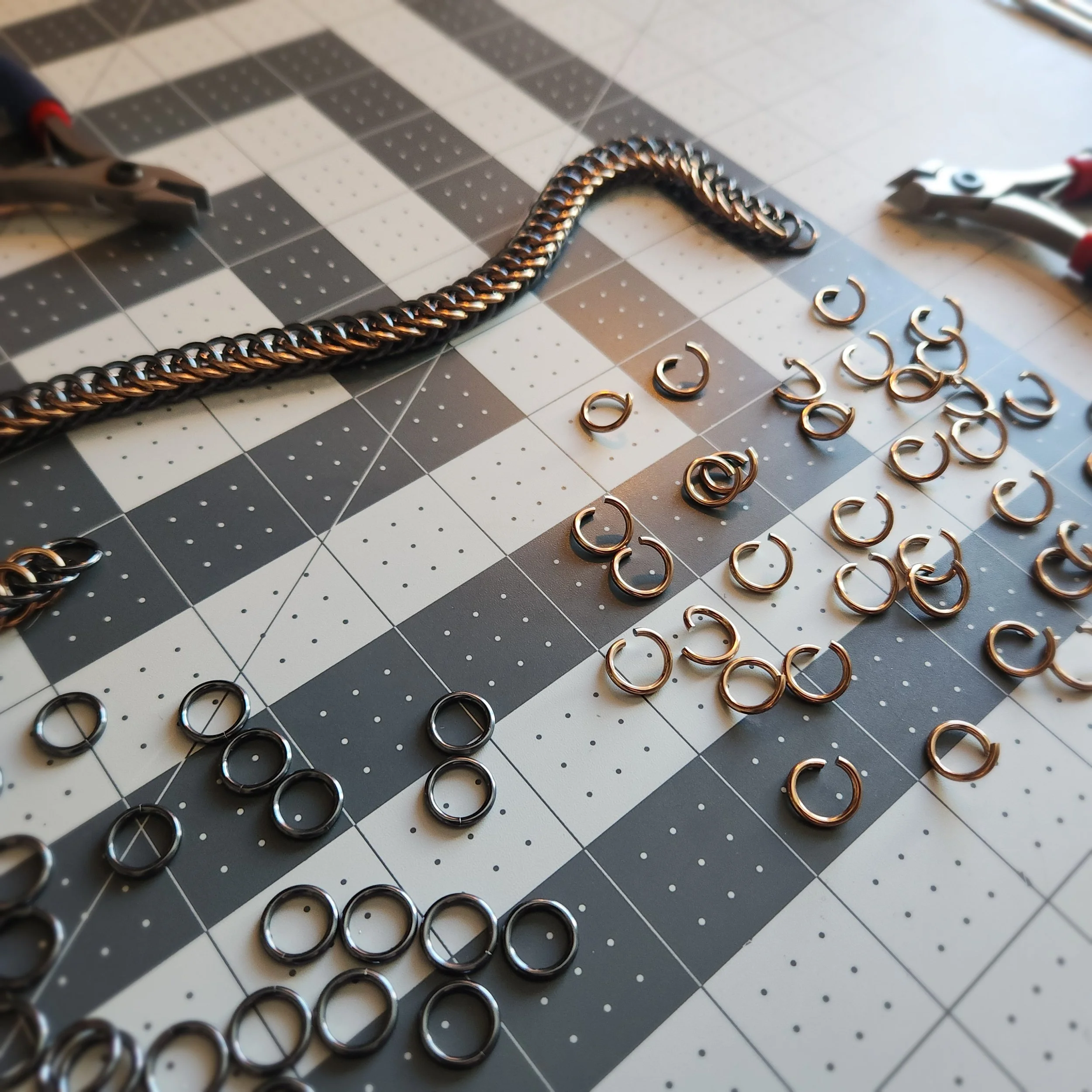 Assorted metal jewelry components, including small rings, covered with a chain and pliers on a black-and-white checkered crafting mat.
