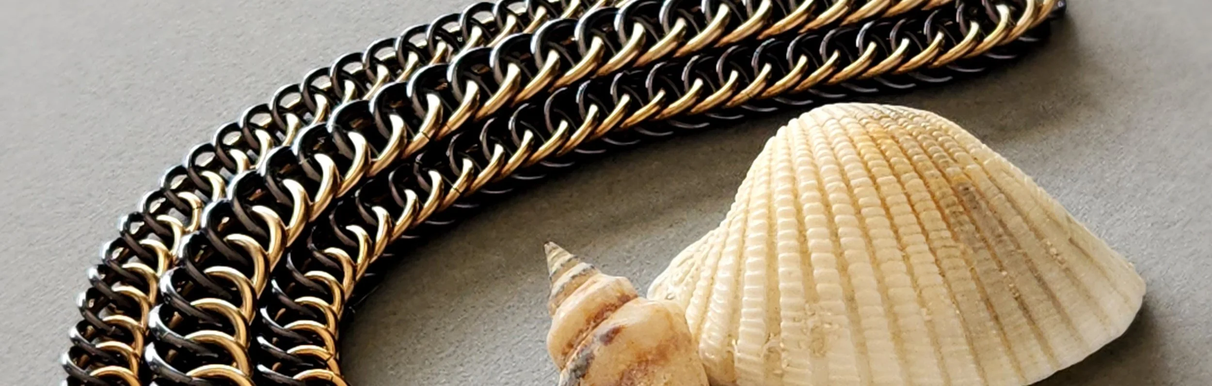 Close-up of three metallic chain necklaces in black, gold, and silver colors, placed on a gray surface with two seashells nearby.