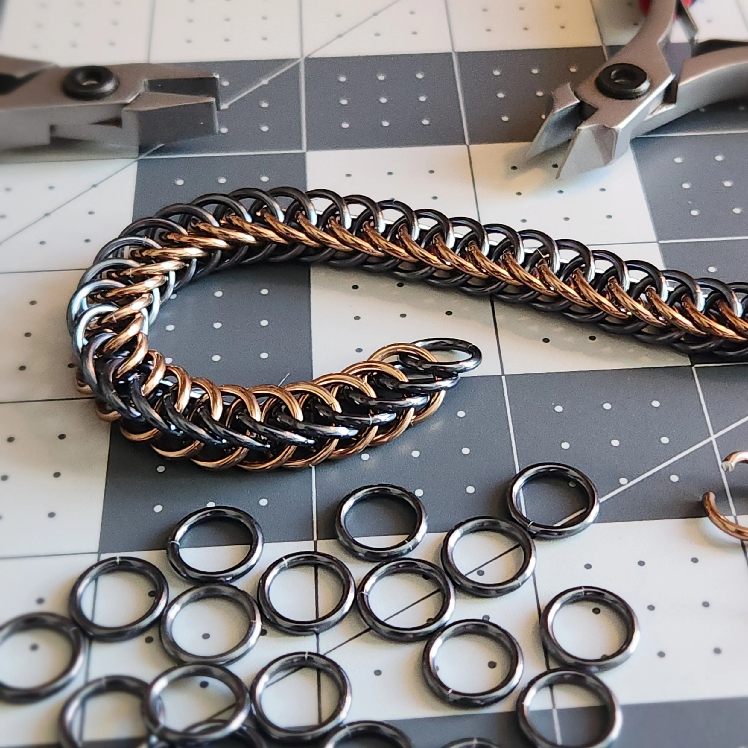 Two braided metal chains on a craft cutting mat, with loose metal rings and pliers in the background.