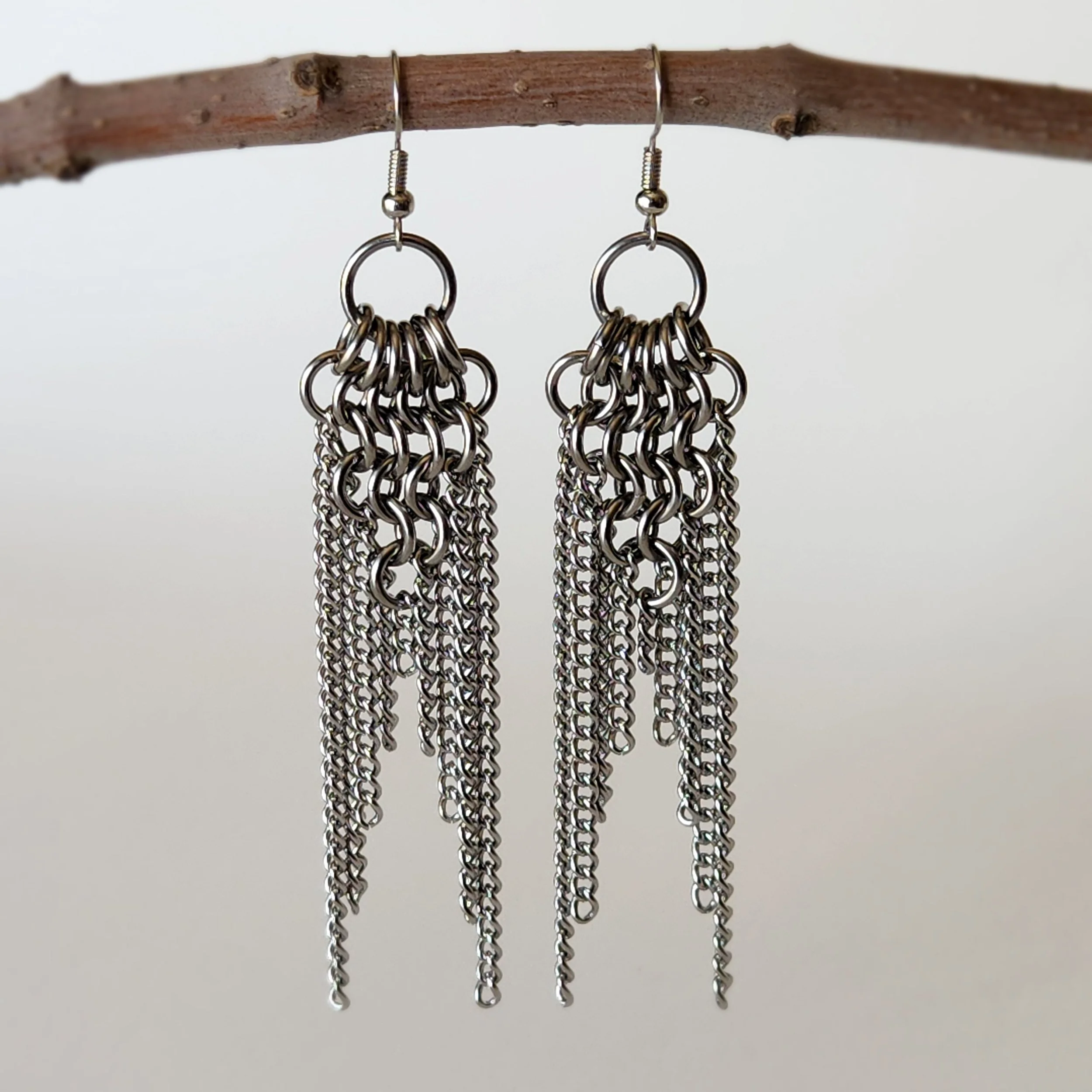 A pair of dangling earrings made of multiple chains hanging from a circular top, suspended from a thin brown twig on a plain background.
