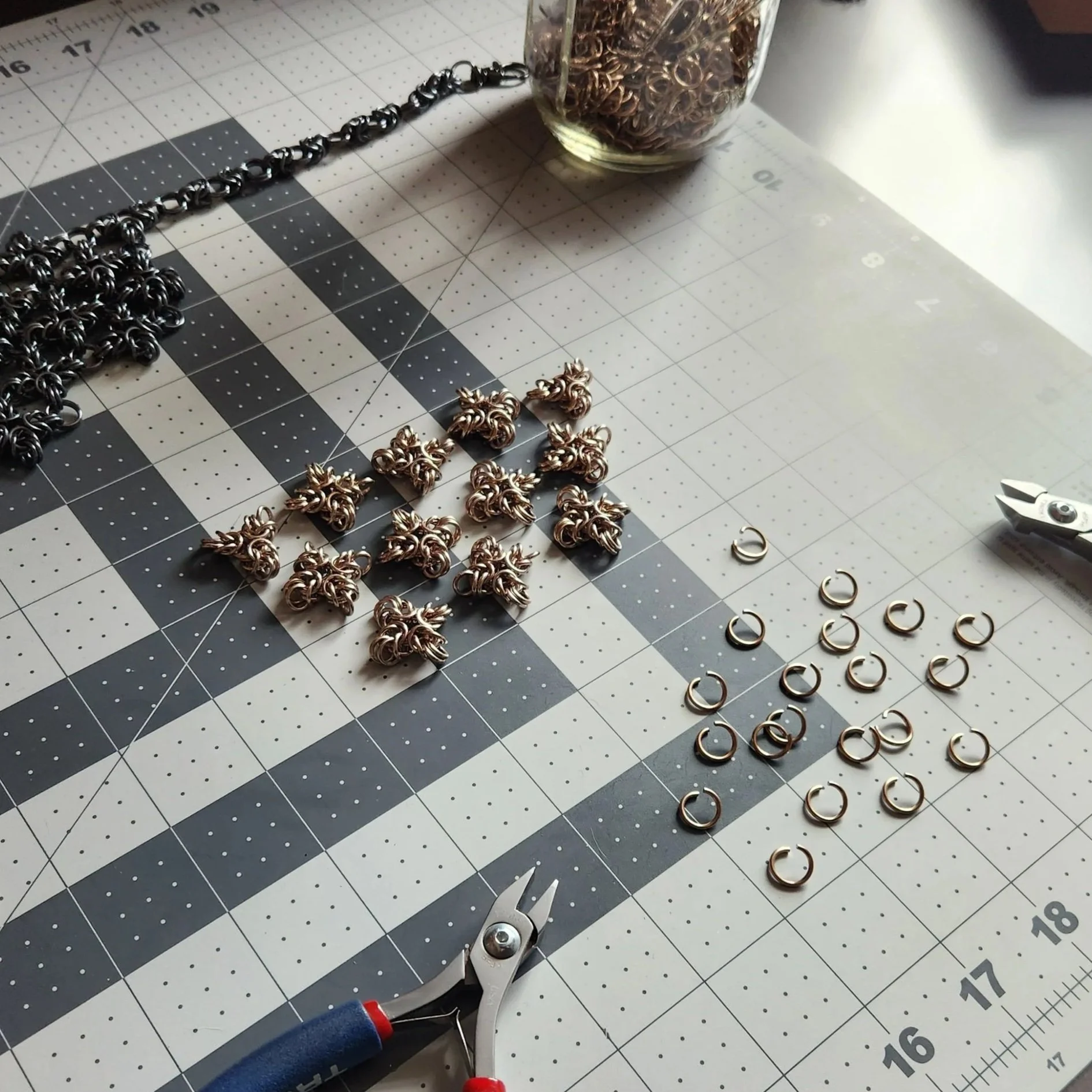 A worktable with jewelry making supplies: a glass jar filled with jump rings, silver chain, various gold jump rings, and gold decorative beads; jewelry pliers; and a cutting mat with measurement markings.
