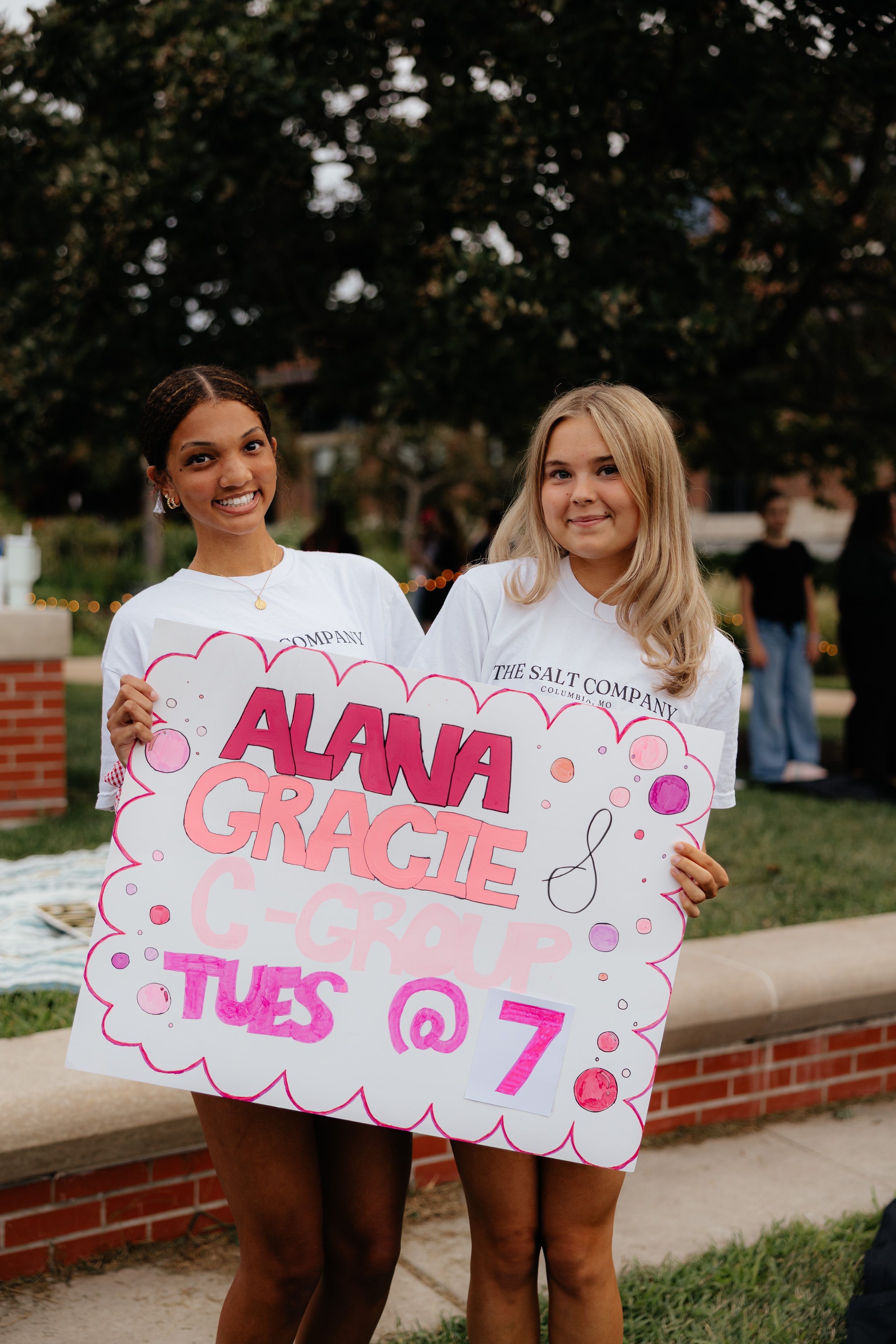 Two young women holding a colorful sign that reads 'Alana & Gracie C-Group Tues @ 7', outdoors with trees and people in the background.