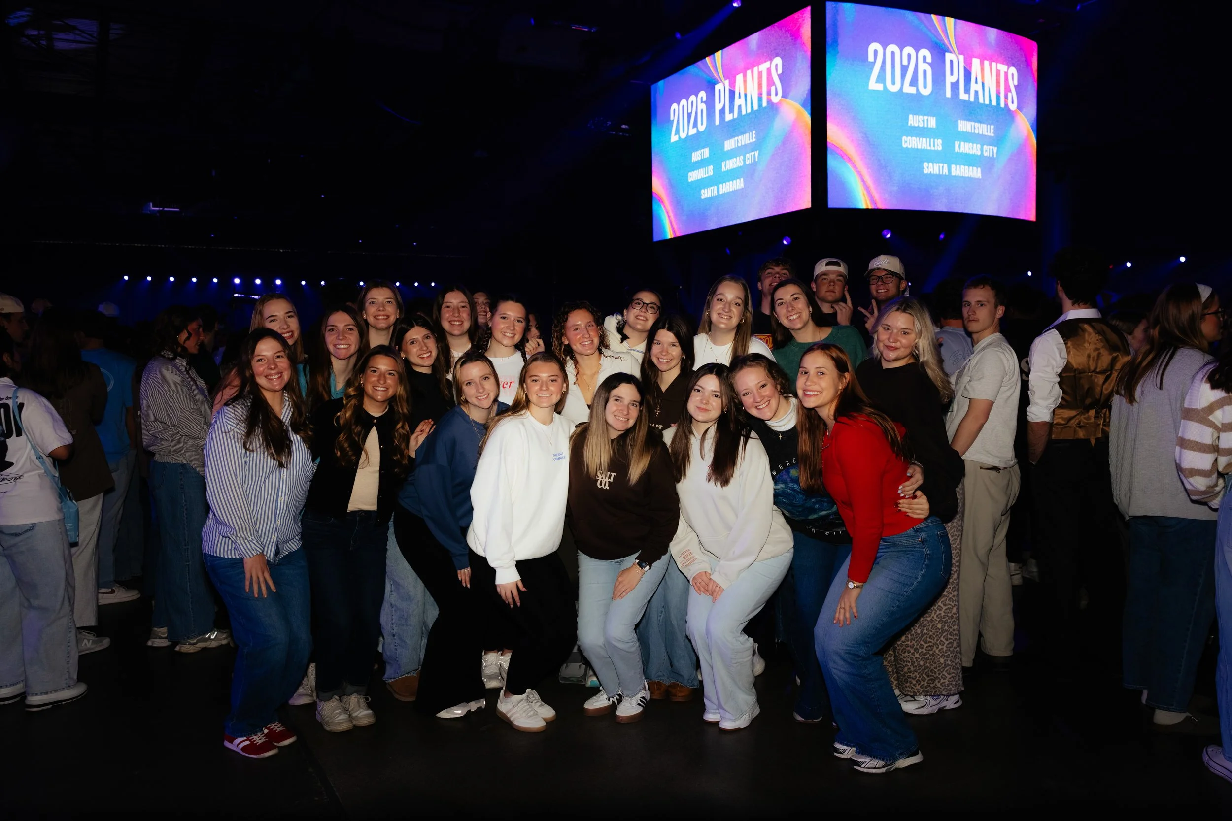 Group of young people smiling and posing for a photo at an event with screens displaying '2026 Church Plants' in the background.