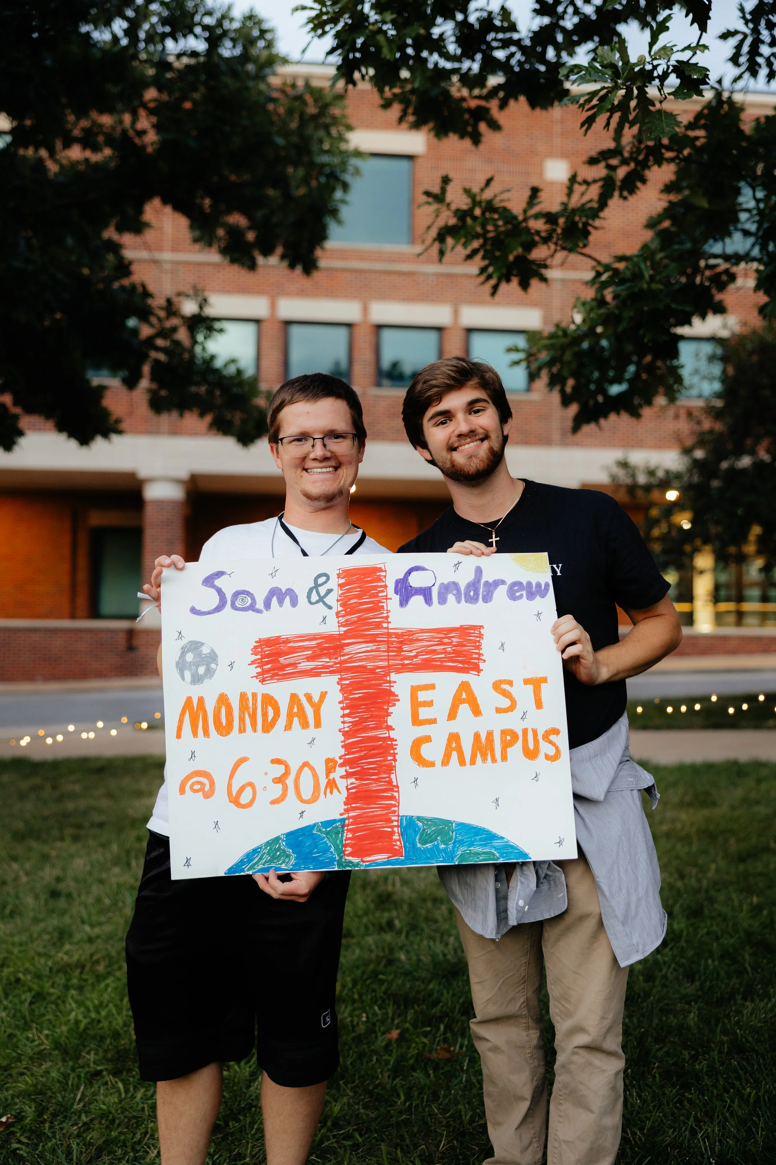 Two young men standing outside on grass holding a colorful hand-drawn sign promoting a community group on Monday at 6:30 PM on East Campus.