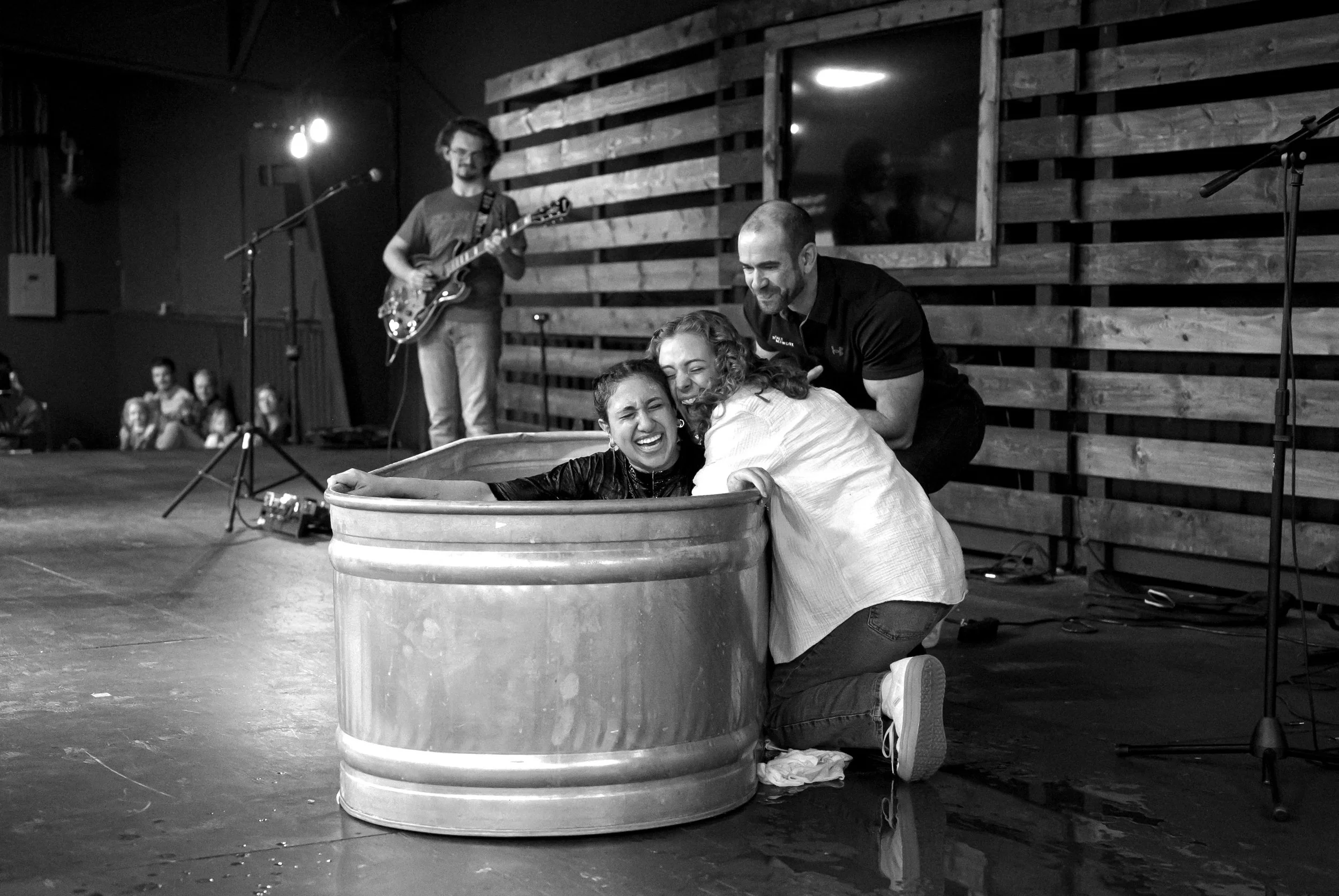 Three people laughing and embracing after a baptism, with a musician playing guitar in the background during a live performance.