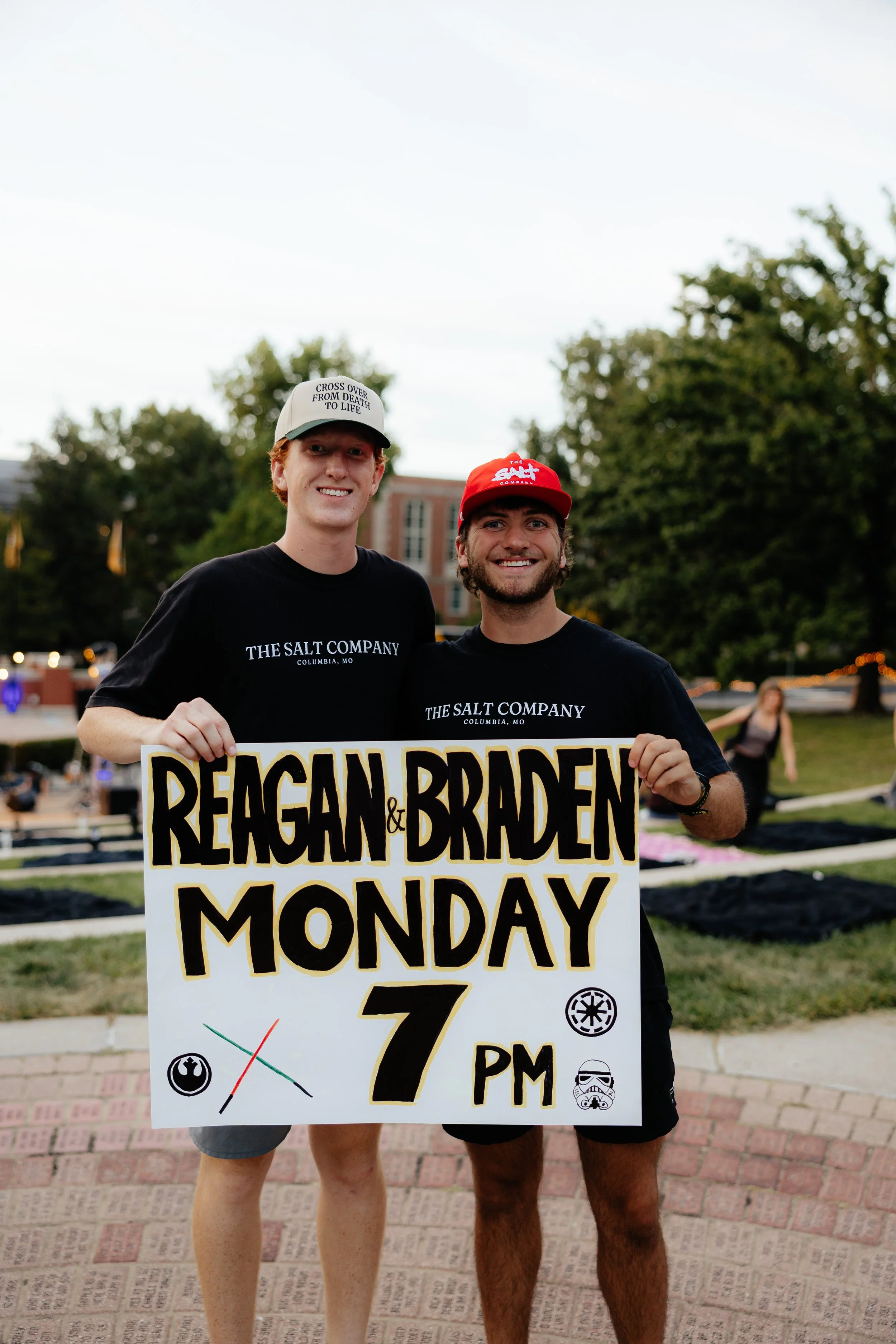 Two young men standing outdoors holding a sign that reads "REAGAN & BRADEN MONDAY 7 PM." They are smiling and wearing black T-shirts labeled "THE SALT COMPANY." The man on the left is wearing a white cap with text, and the man on the right is wearing a red cap. The background shows a park or public space with people and trees.