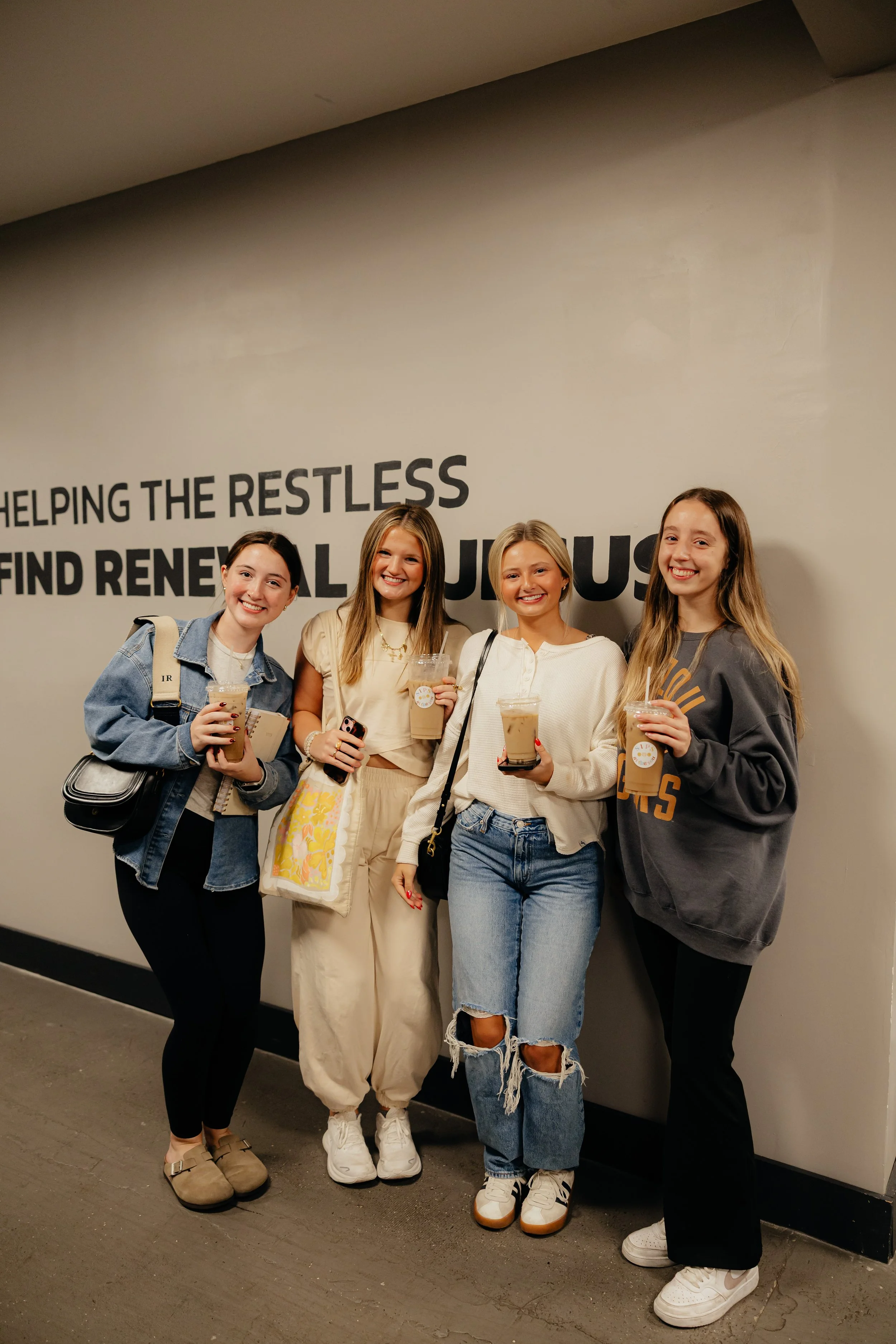 Four young women standing together, smiling, holding iced coffee drinks, and posing in front of a wall with inspiring text.