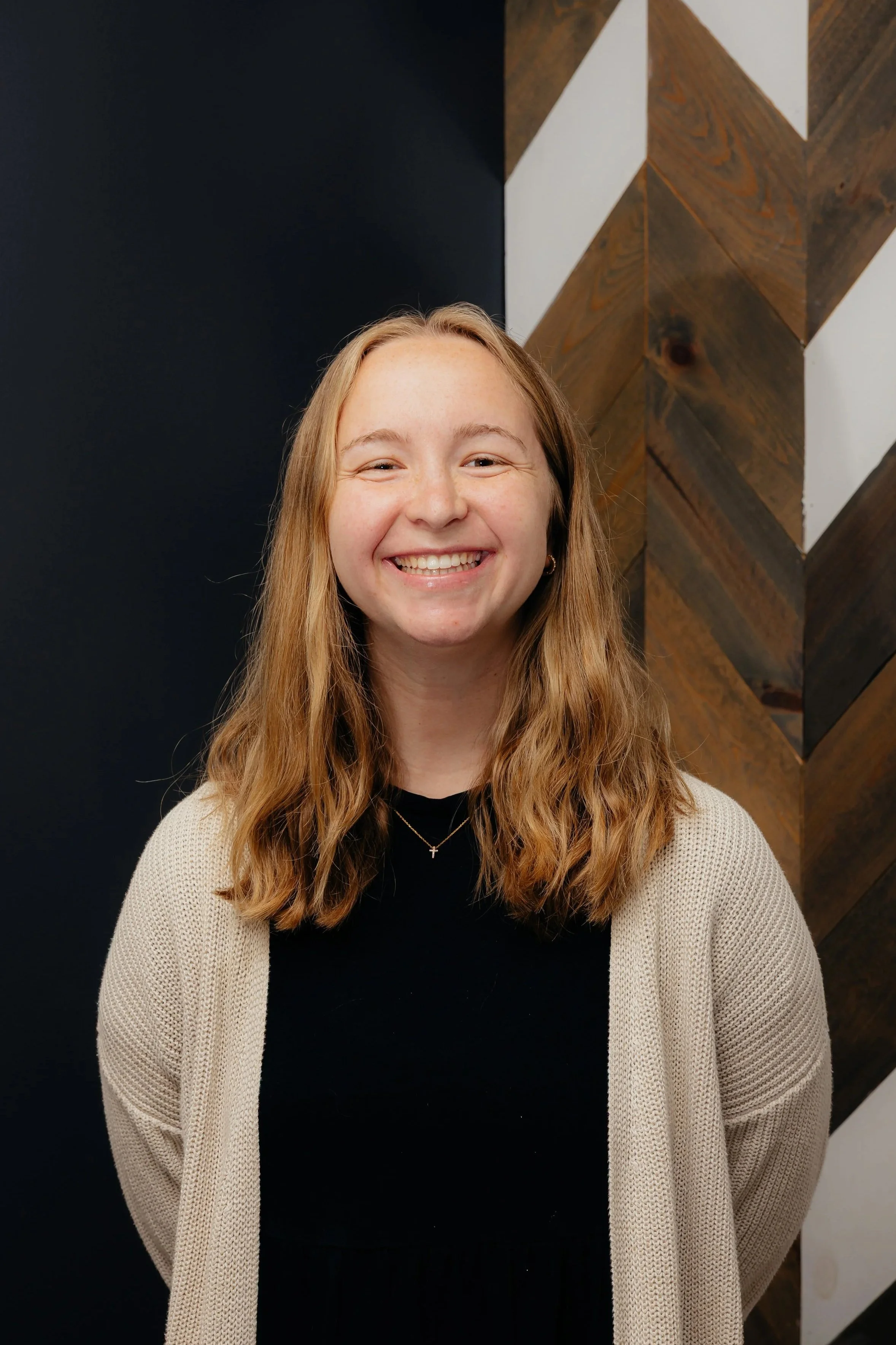 A young woman with blonde hair smiling at the camera, standing in front of a wall with a chevron pattern in the background.