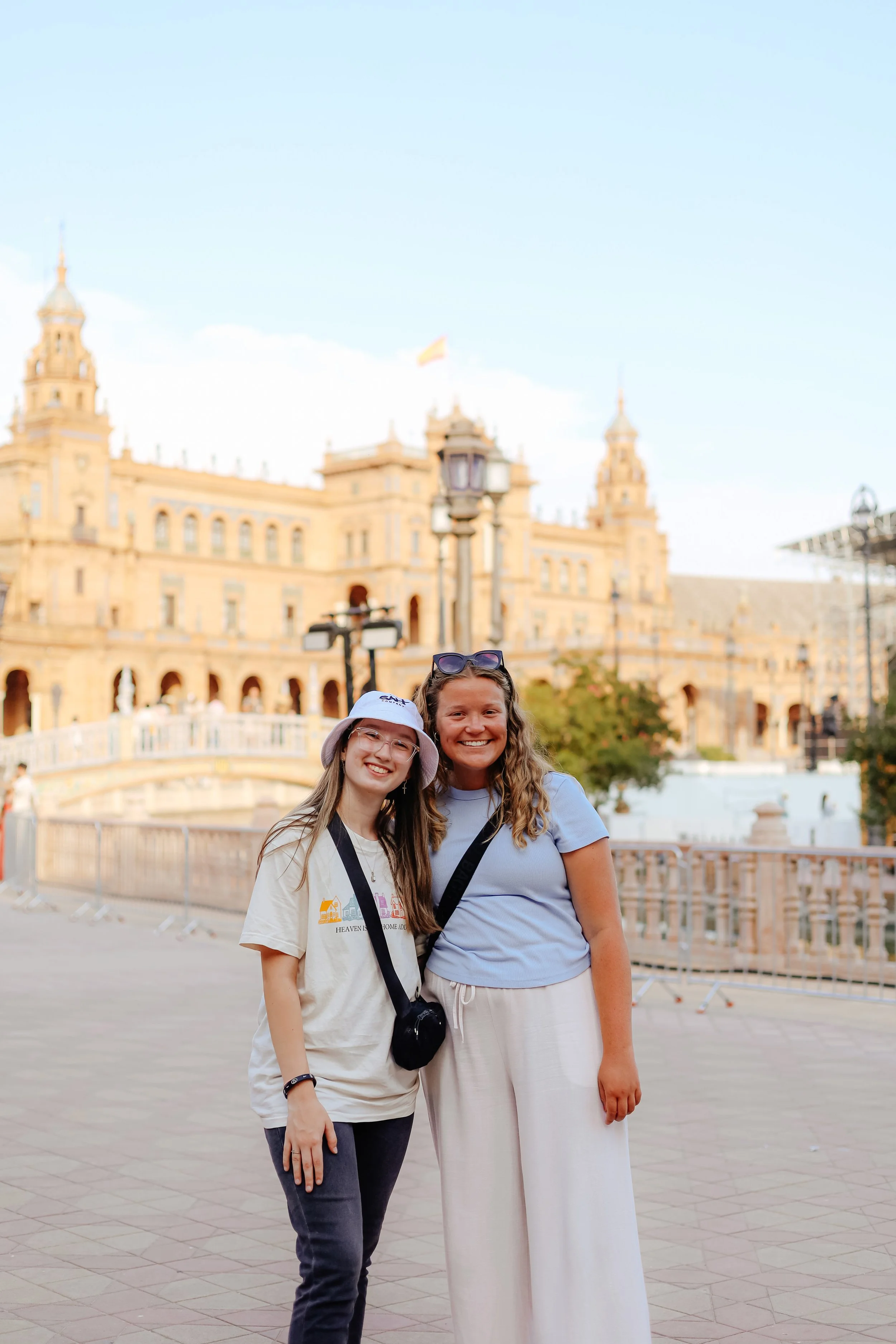 Two young women smiling and standing together outdoors in front of a historic castle, with clear blue skies above.
