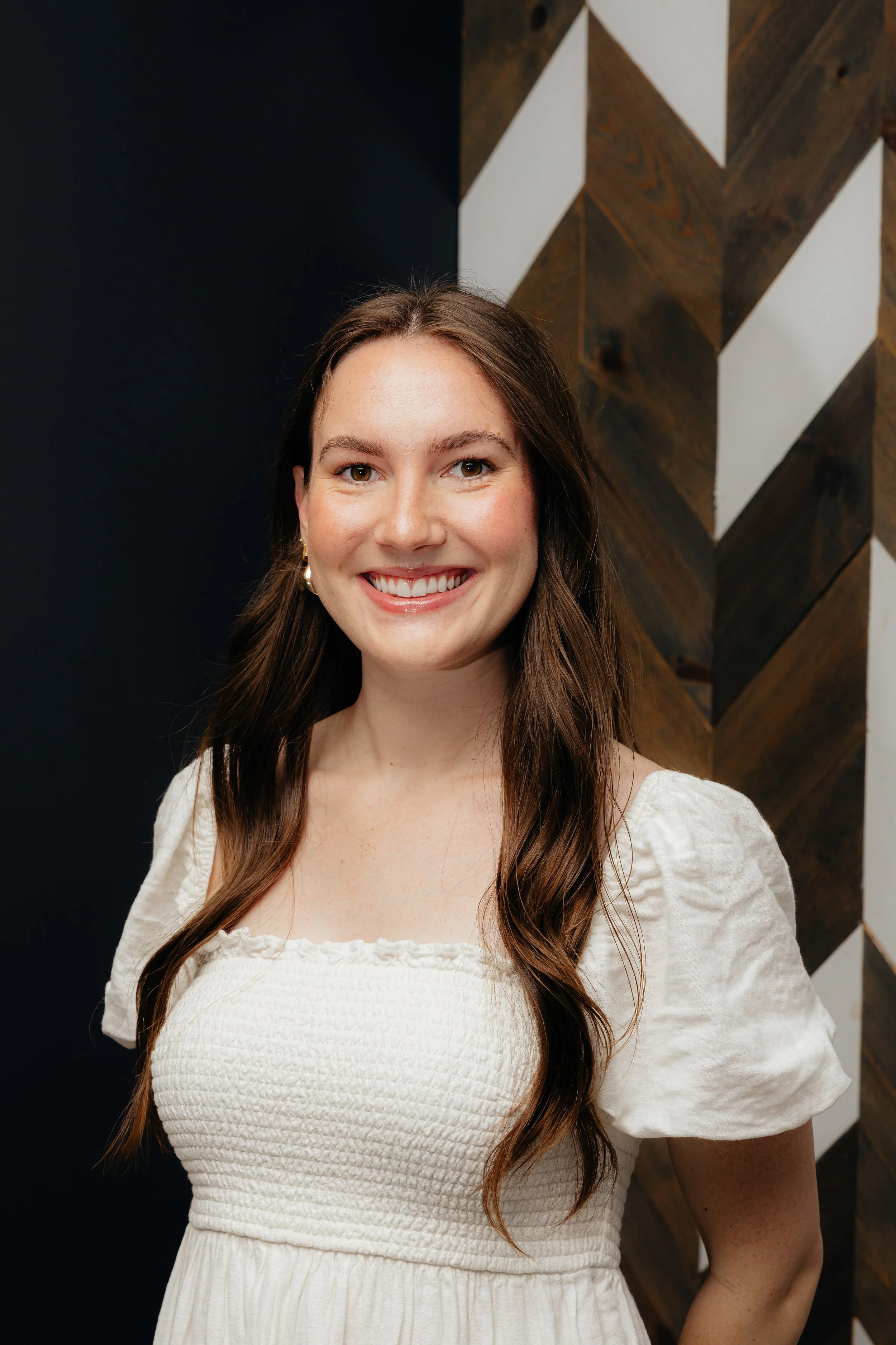 A smiling woman with long brown hair wearing a white dress standing in front of a dark wall with wooden and white geometric design.