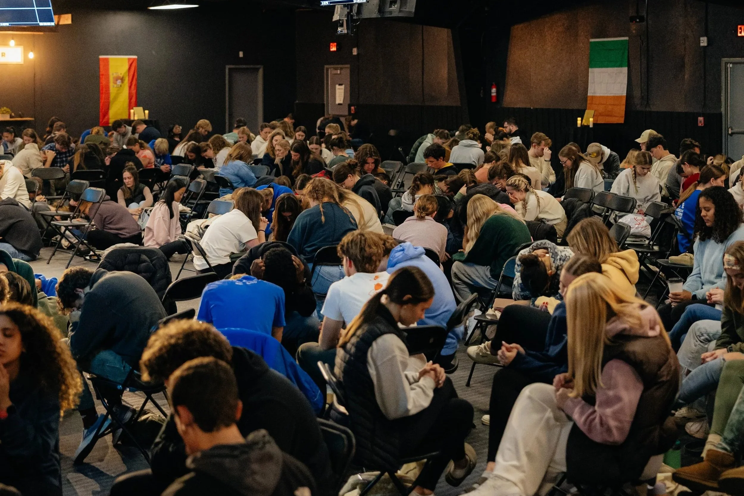A large group of people sitting and kneeling on chairs and the floor in a dark room, with flags of Spain and Ireland hanging on the back wall, indicating a multicultural or international event.