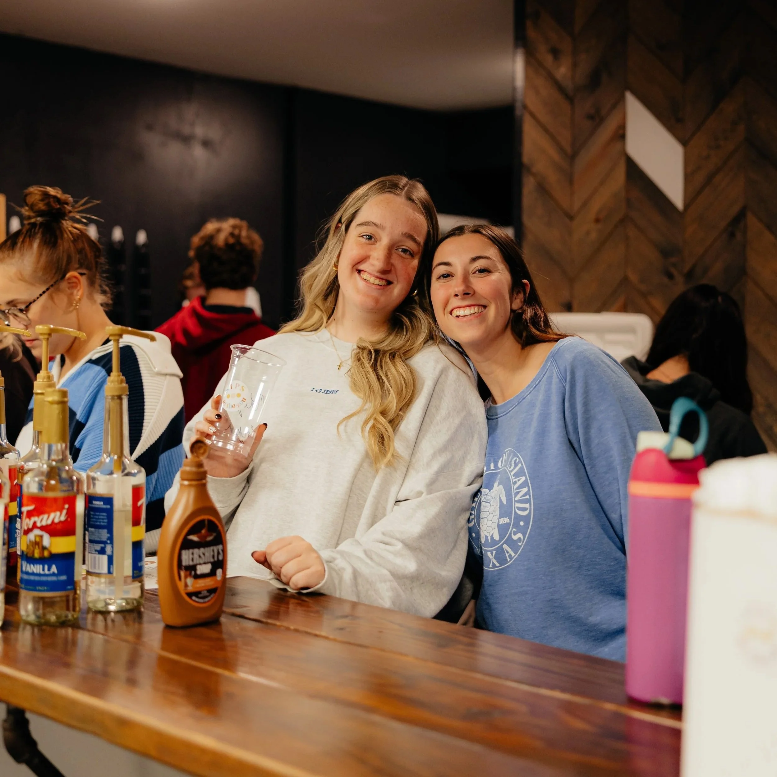 Two young women smiling and leaning close together at a wooden table, with drinks, syrups, and personal items on the table in a cozy indoor setting.