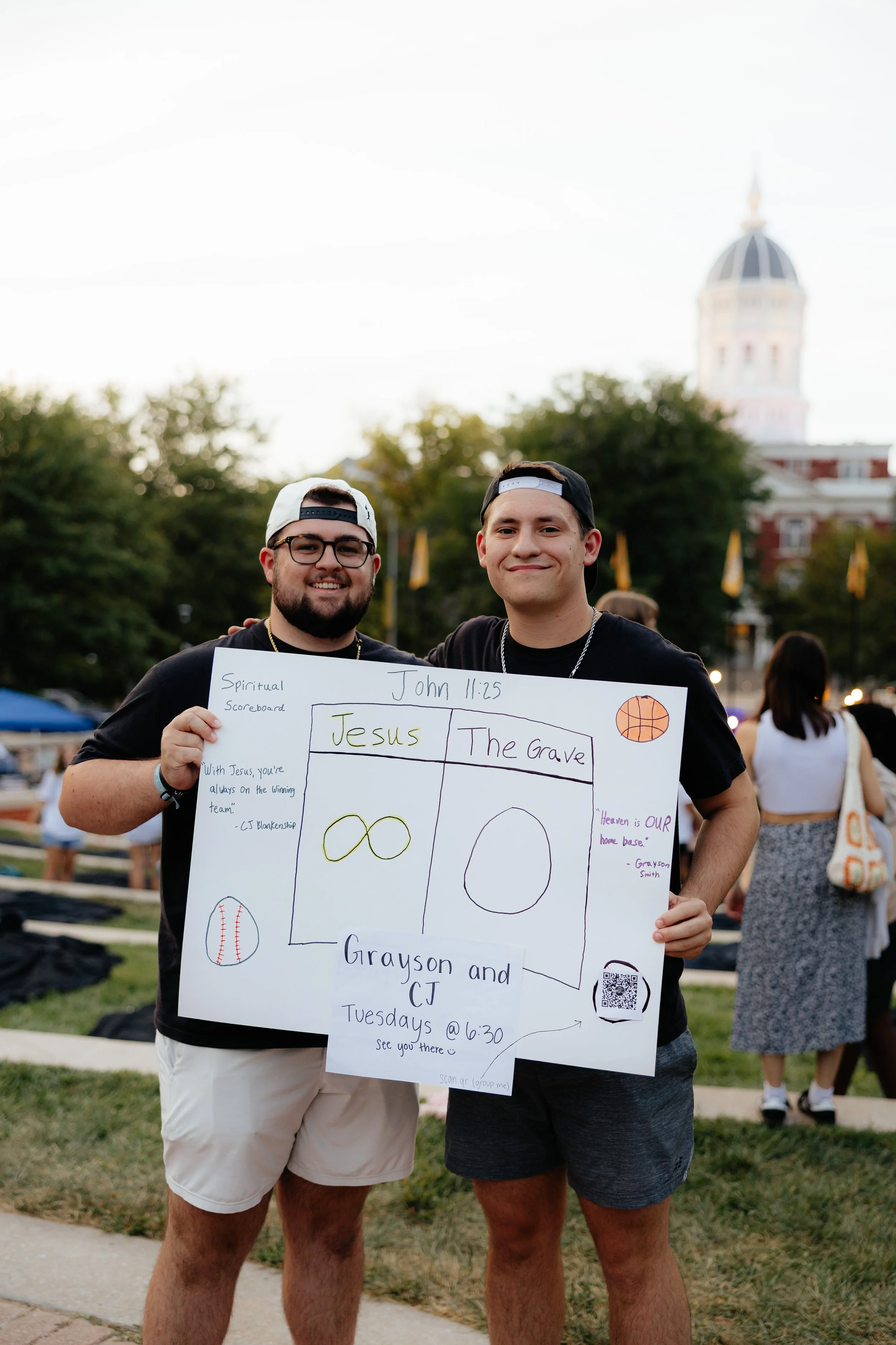 Two young men standing outdoors on a grassy area, holding a large poster board with handwritten notes about c-group.