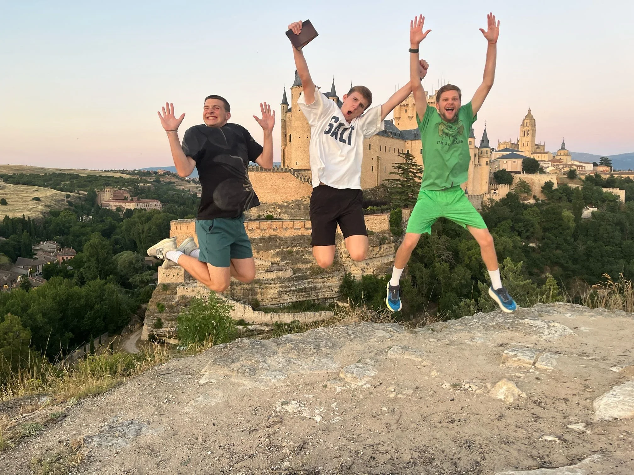 Three young men jumping in the air with a castle in the background, during sunset, on a rocky terrain.