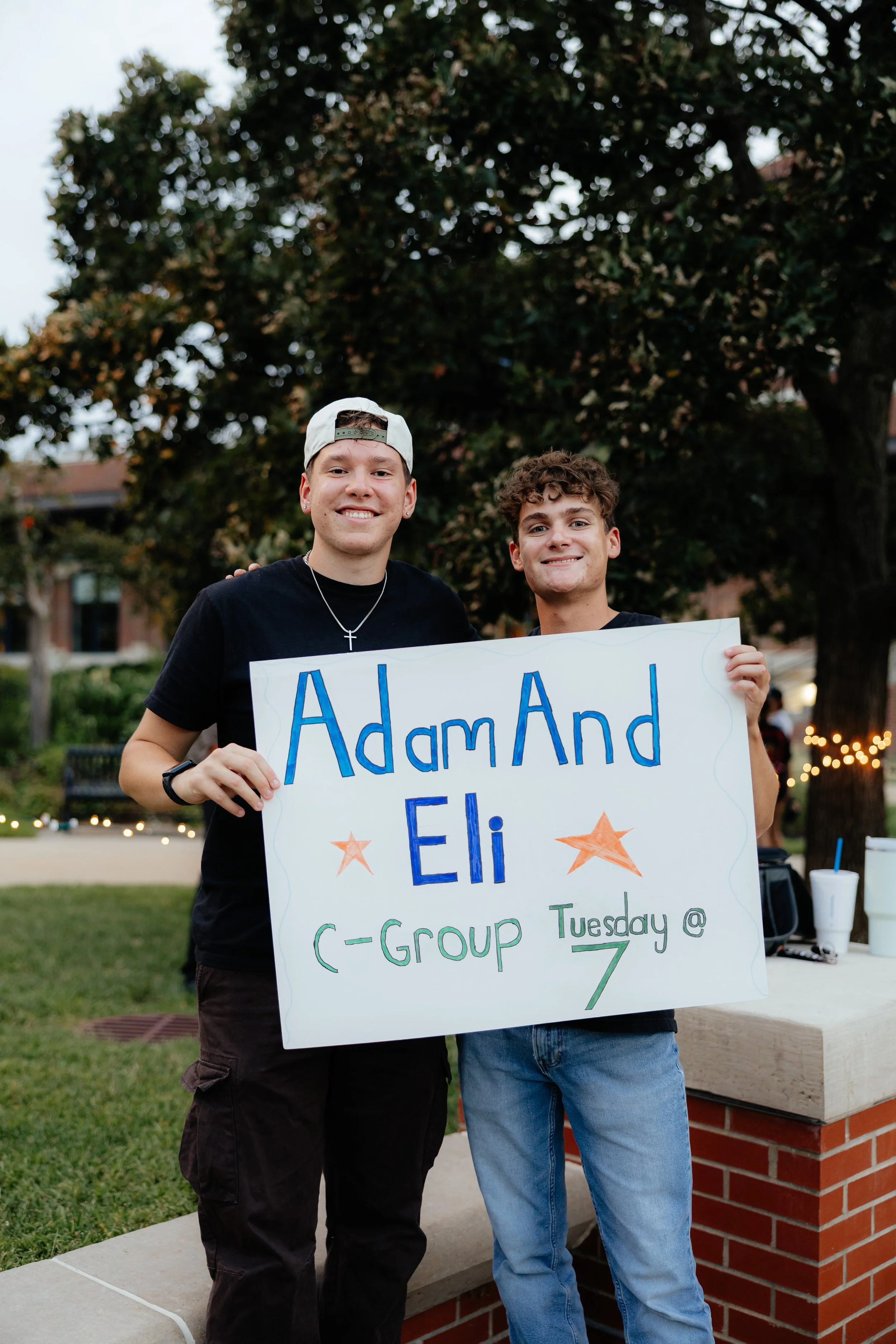 Two young men smiling and holding a large sign that says 'Adam and Eli C-Group Tuesday @ 7' outdoors during the evening with trees and string lights in the background.