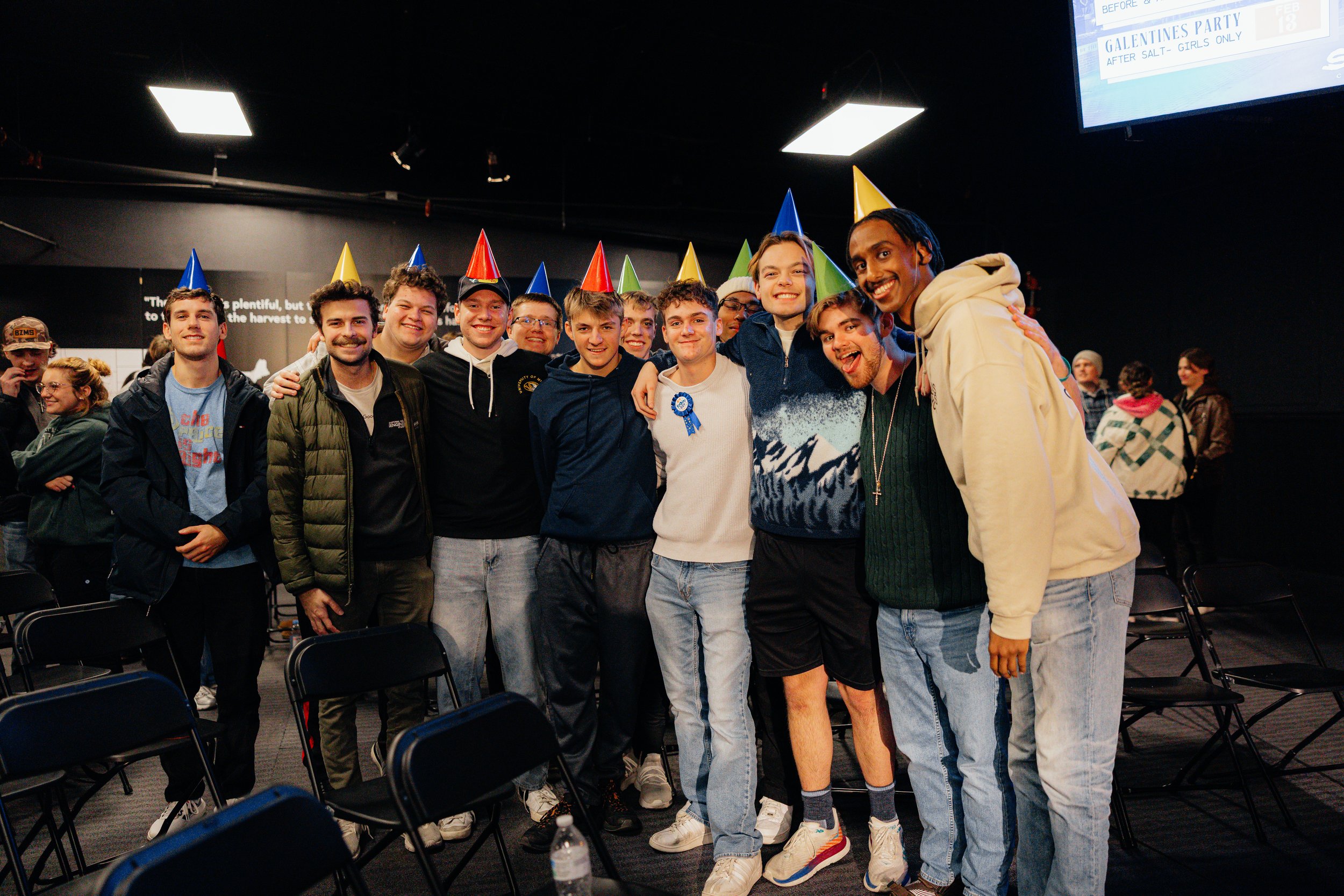 Group of young men wearing colorful party hats, smiling and standing close together at a celebration event.