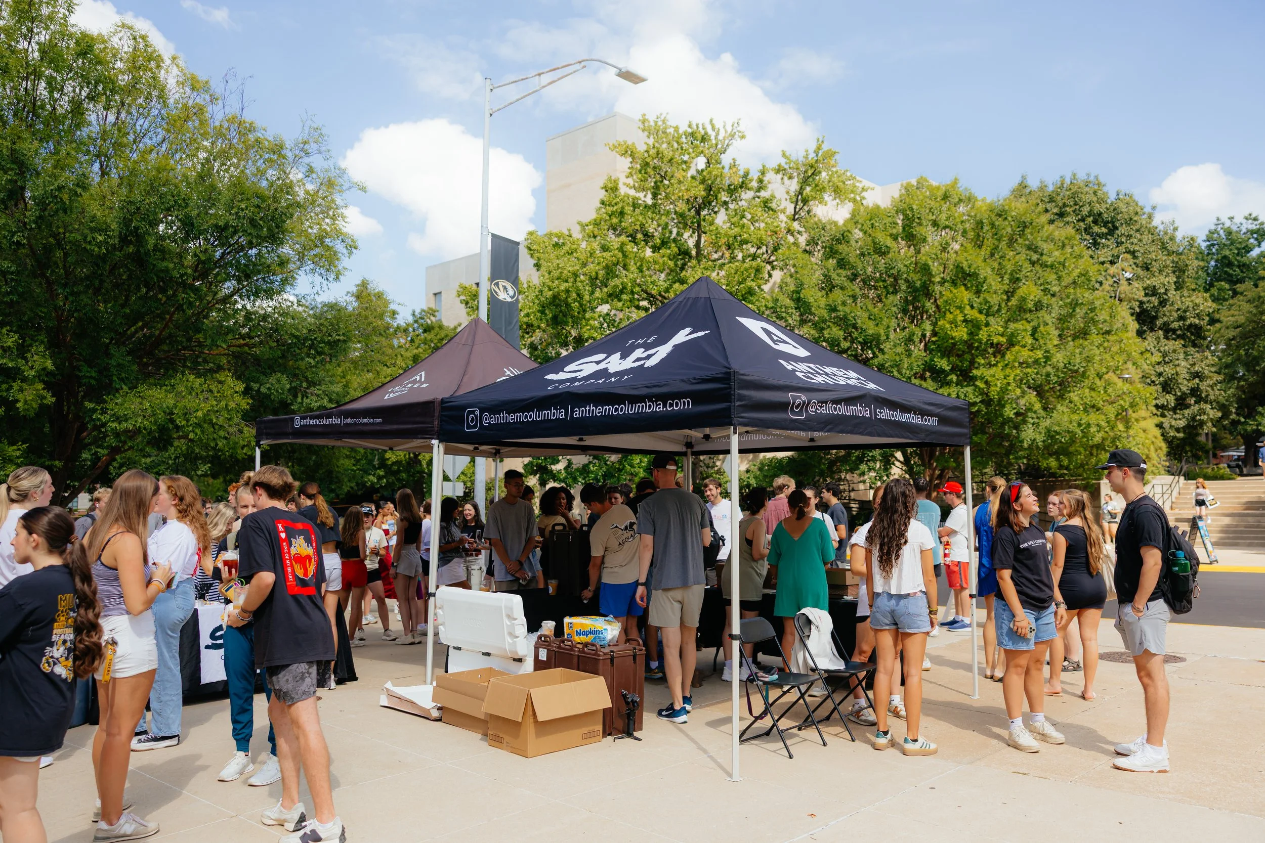 Crowd of people gathered around a booth on a sunny day, with trees and buildings in the background.