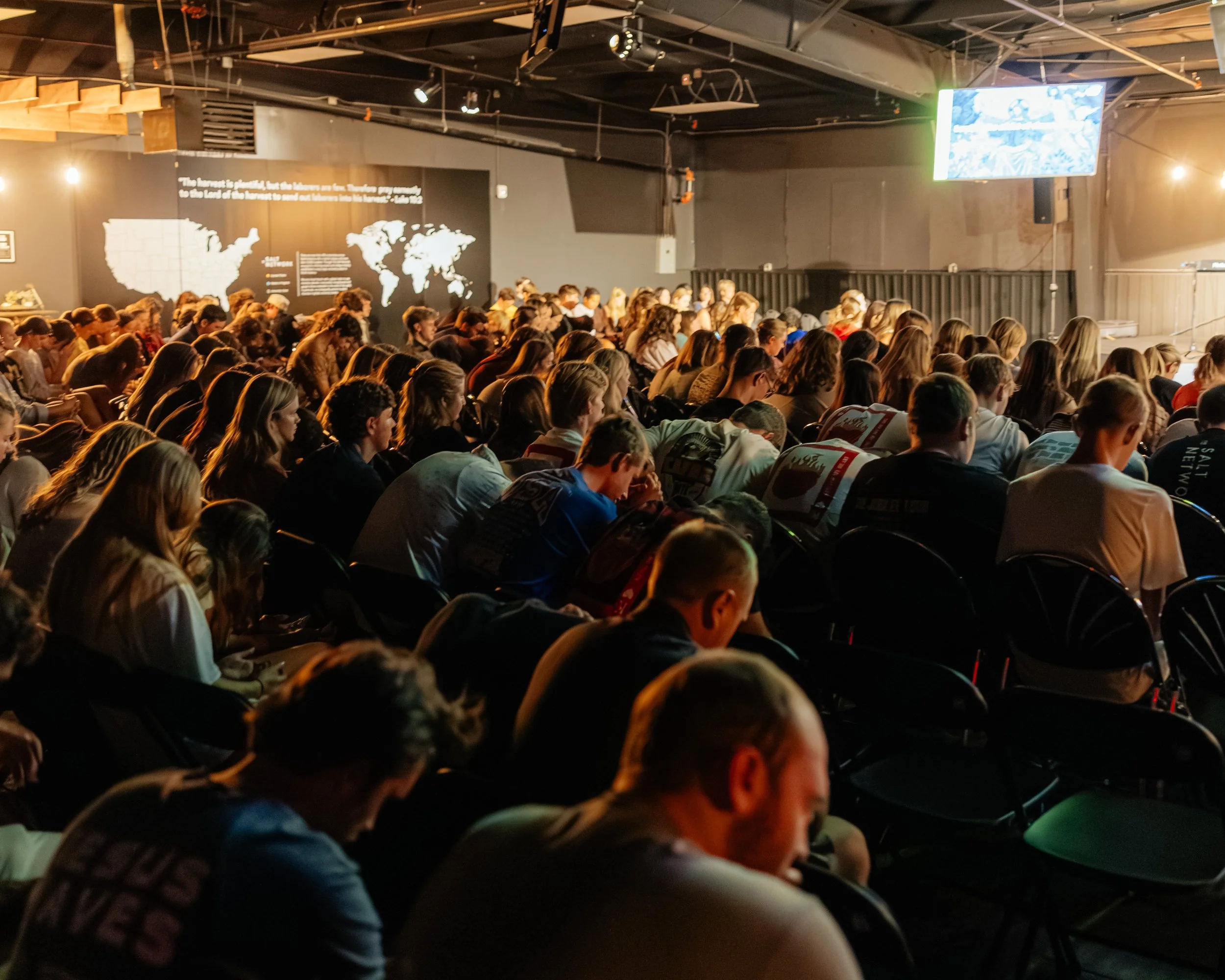 A large group of people sitting in a dark auditorium, audience members are watching a presentation or performance on a stage.