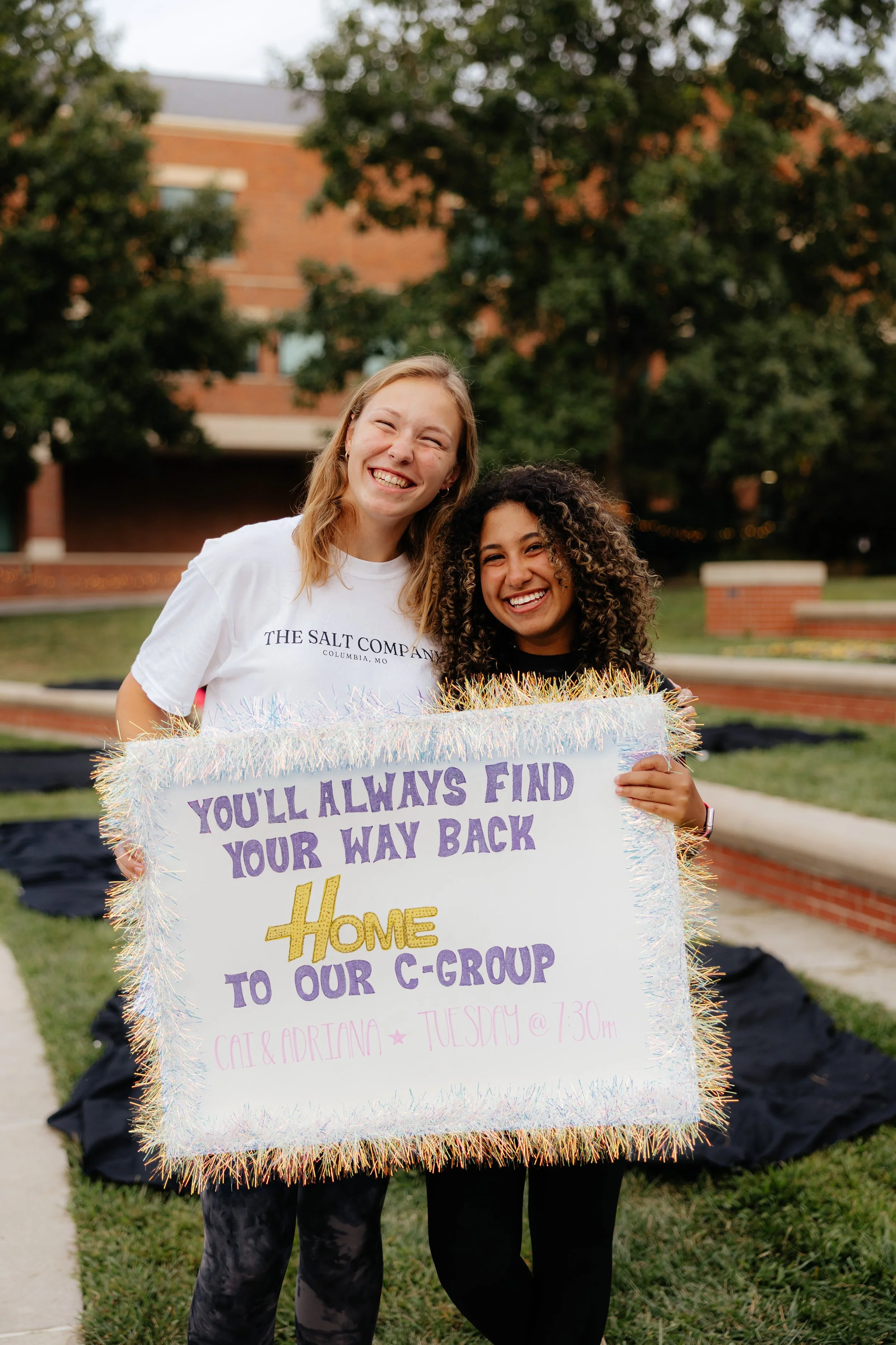 Two women smiling and holding a colorful sign that says, 'You'll always find your way back home to our C-group' at an outdoor setting with trees and a brick building in the background.