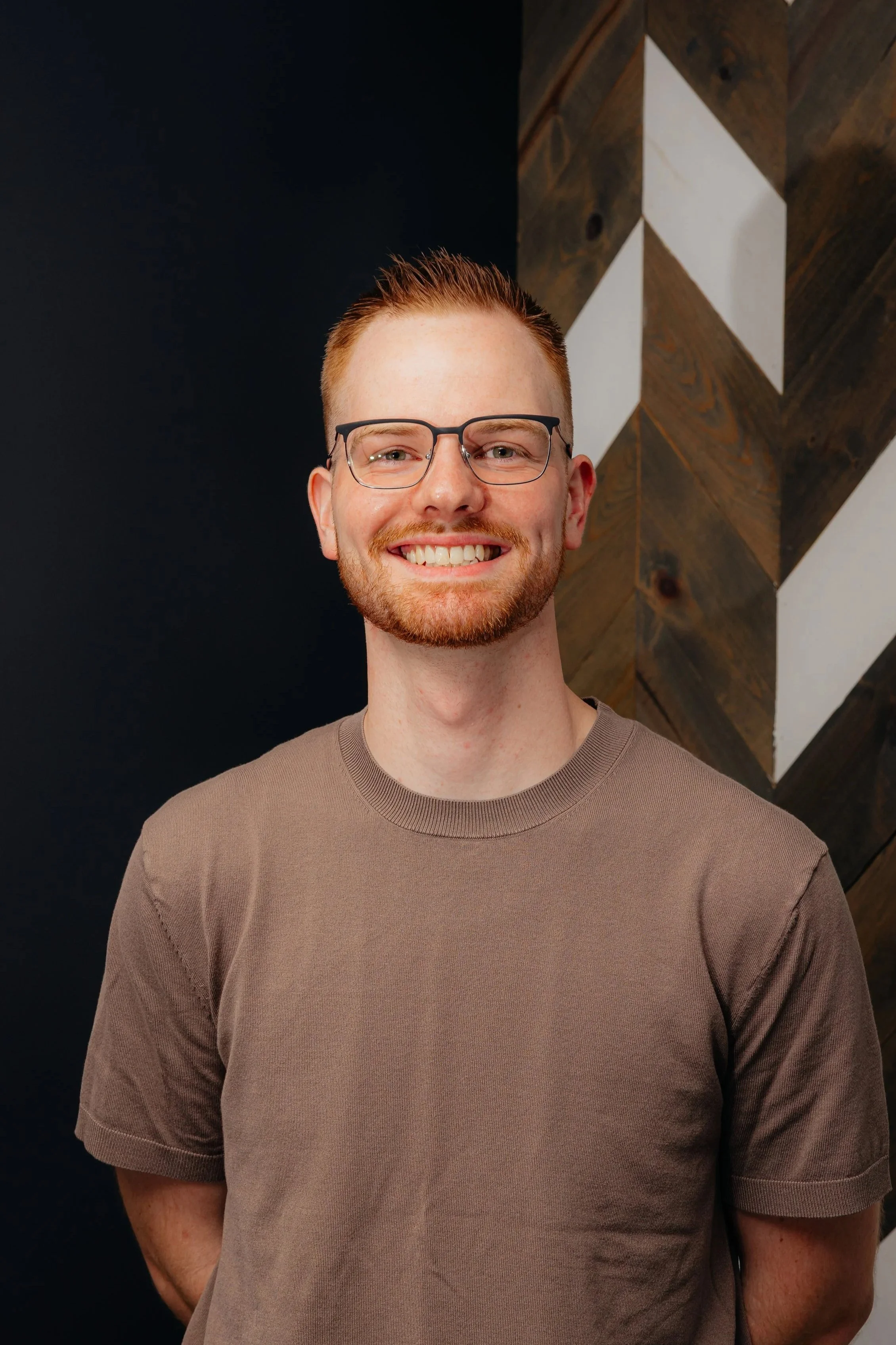 A smiling young man with short red hair, beard, glasses, and wearing a brown t-shirt stands in front of a black and wood textured background.
