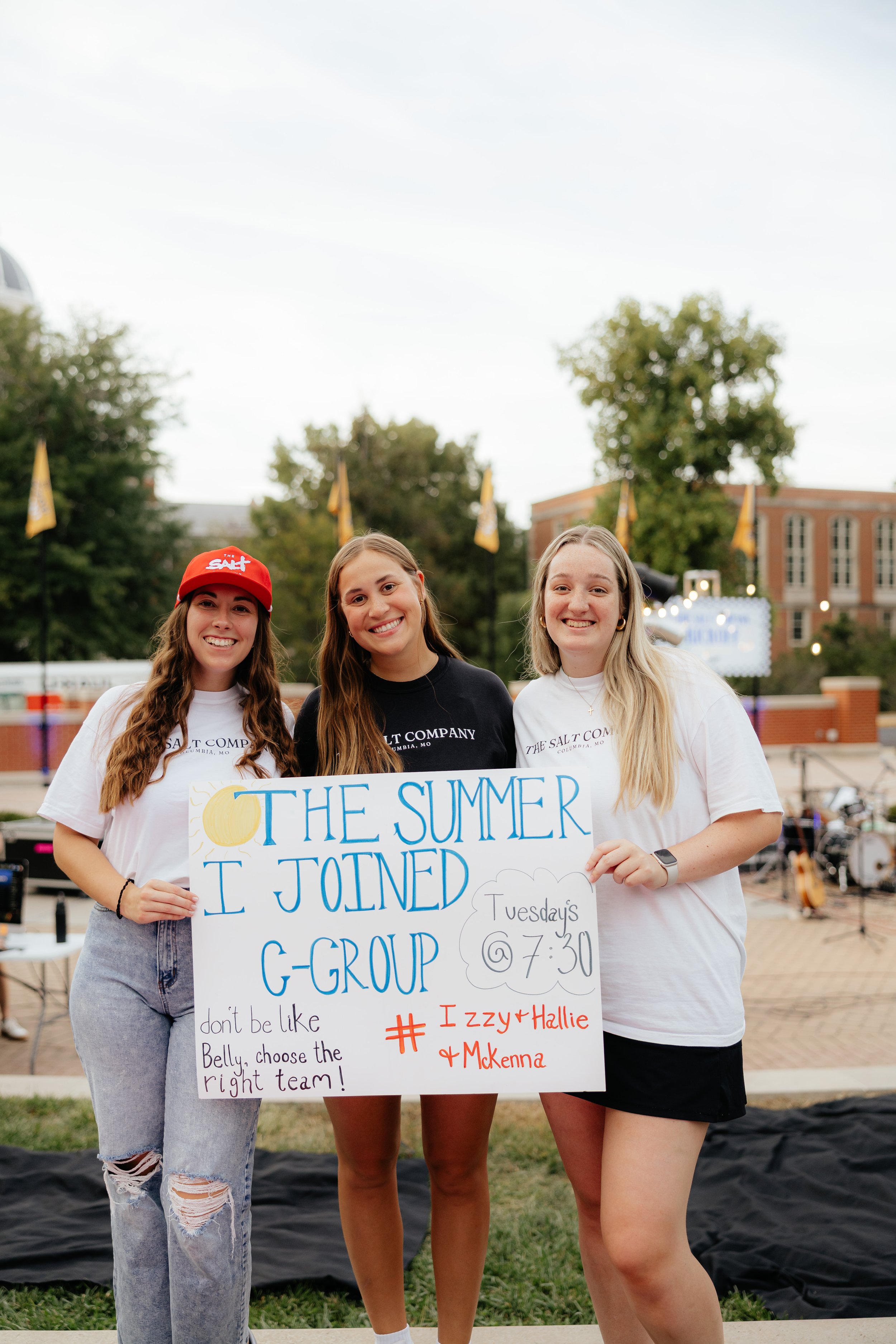 Three young women smiling and holding a sign that reads "The Summer I Joined G-Group". The sign also says "Tuesdays @ 7:30" and has a note "don't be like Belly, choose the right team!" along with "#Izzy+Hallie+McKenna" written on it.