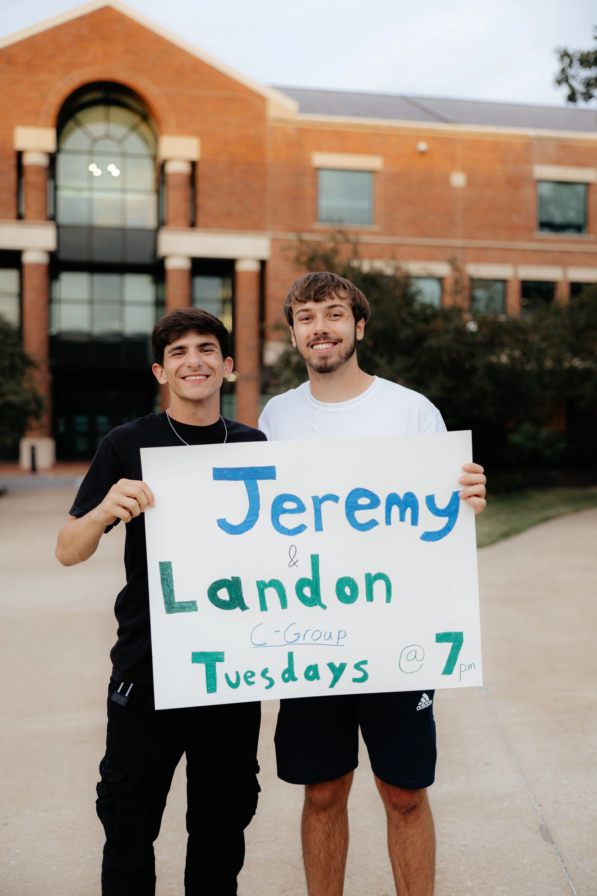 Two young men smiling outdoors, holding a sign that reads 'Jeremy & Landon C-Group Tuesdays @7 pm', in front of a brick building with large windows.