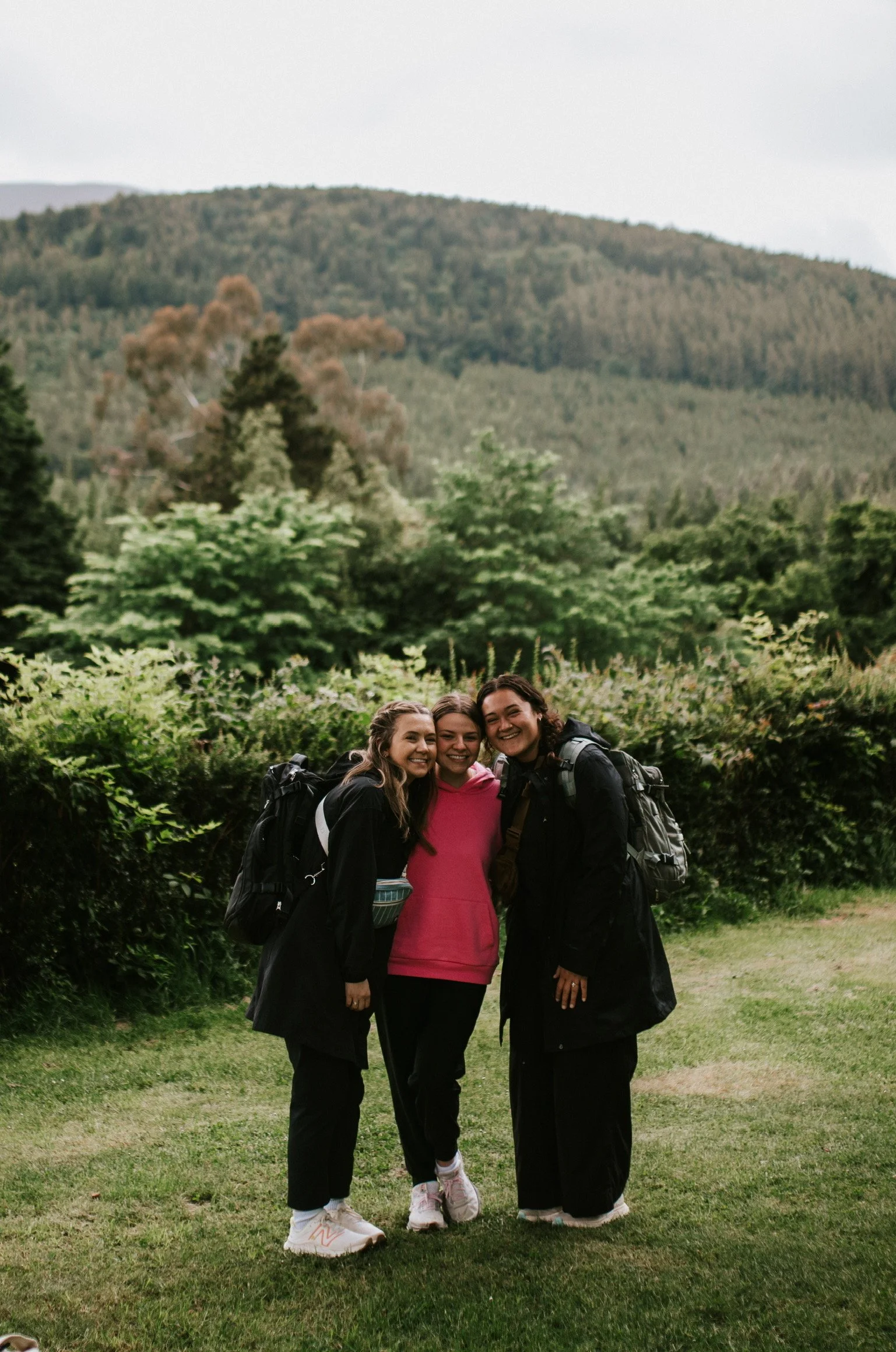 Three women standing outdoors on a grassy area with green bushes and trees, smiling together. They are dressed in casual outdoor clothing with backpacks, and a forested hillside is in the background.