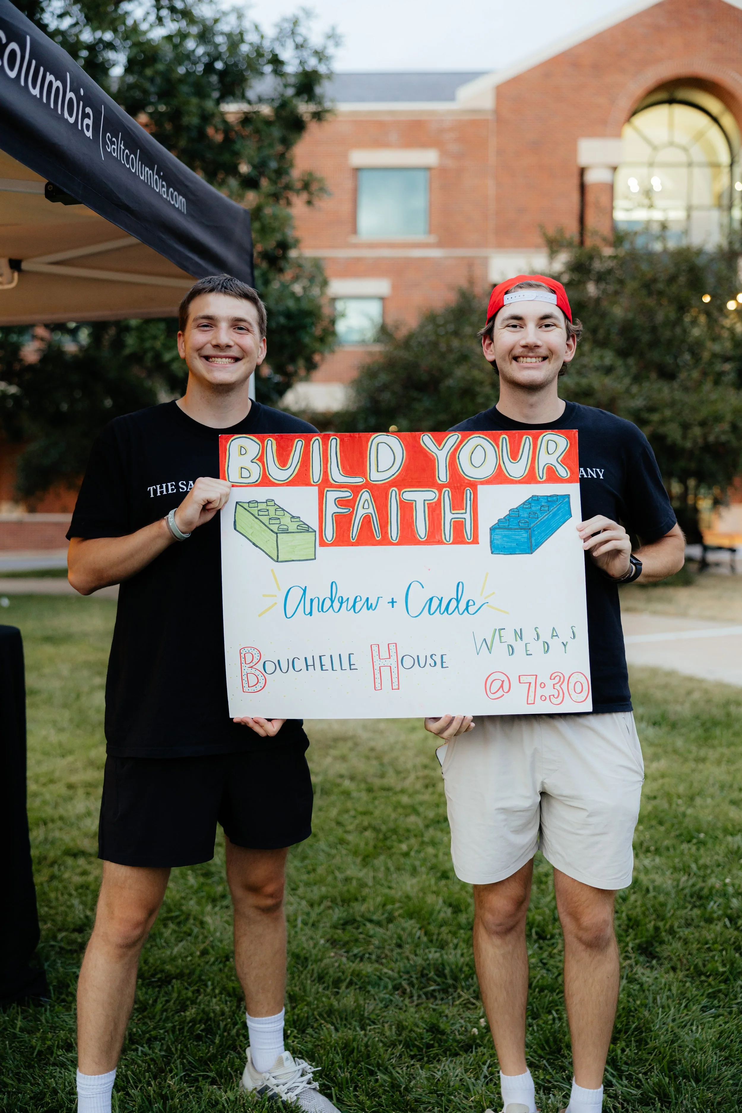 Two smiling young men standing outdoors on grass, holding a colorful hand-made sign that reads 'Build Your Faith' with illustrations of Lego blocks, and details about an event at Bouchelle House at 7:30, including the names Andrew and Cade.