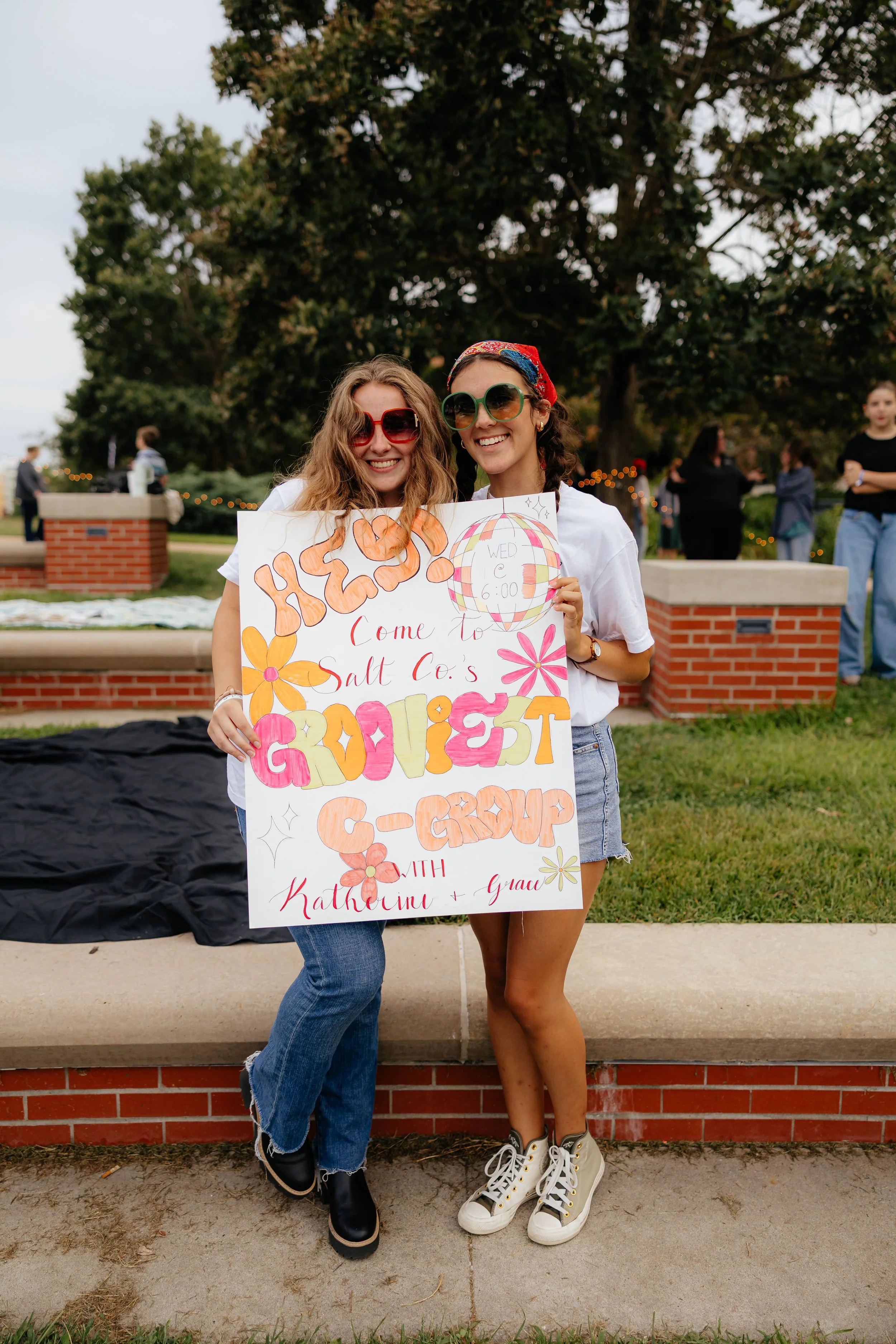 Two women holding a colorful poster at an outdoor event, smiling, with trees and other people in the background.
