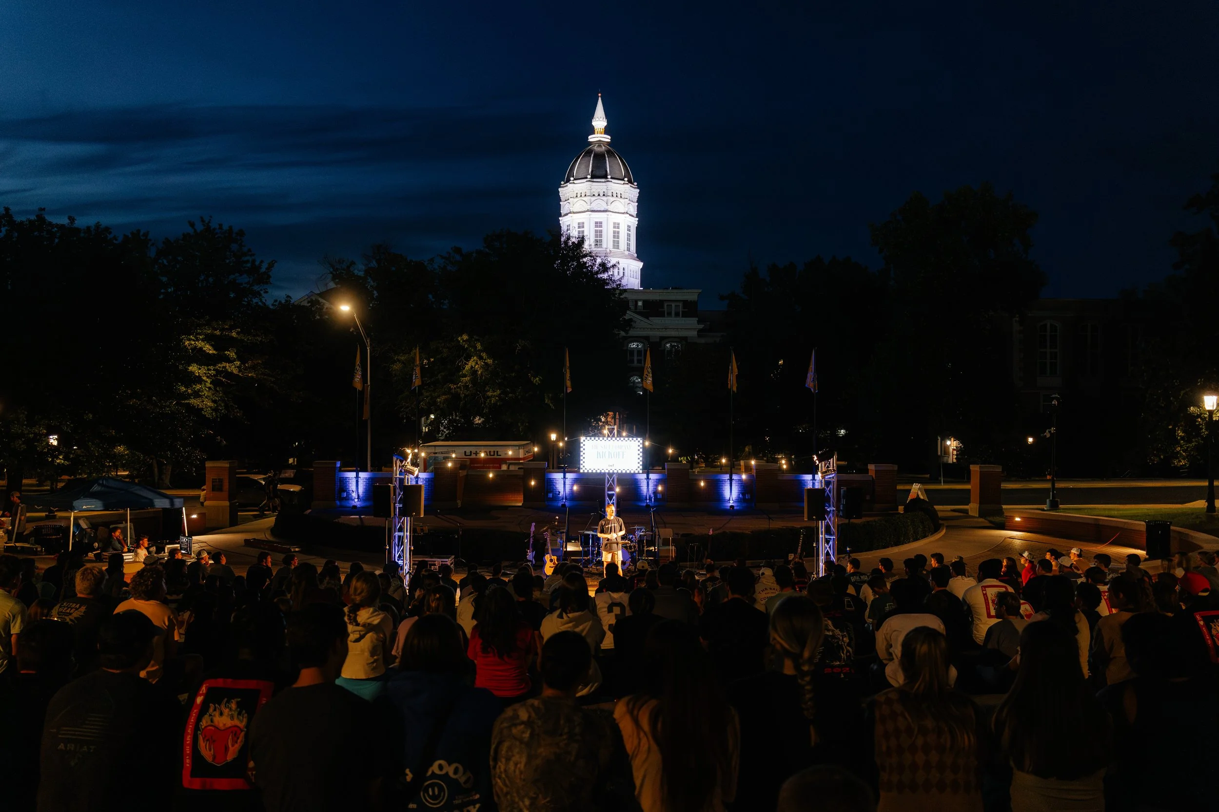 Crowd gathered at an outdoor event at night with a musician performing on stage, illuminated by stage lights.