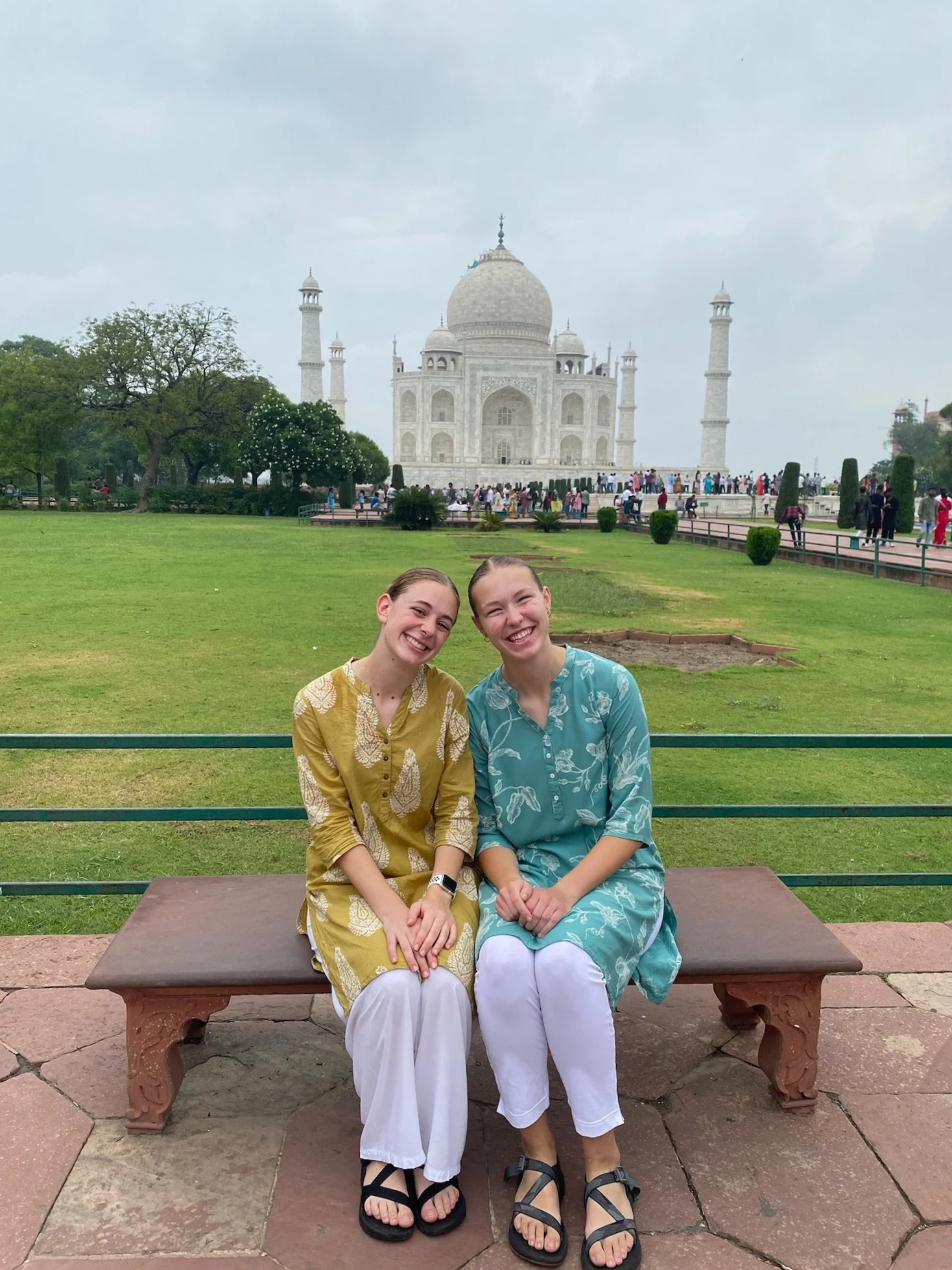 Two women sit on a bench in front of the Taj Mahal, smiling, with the Taj Mahal visible in the background and many people around.