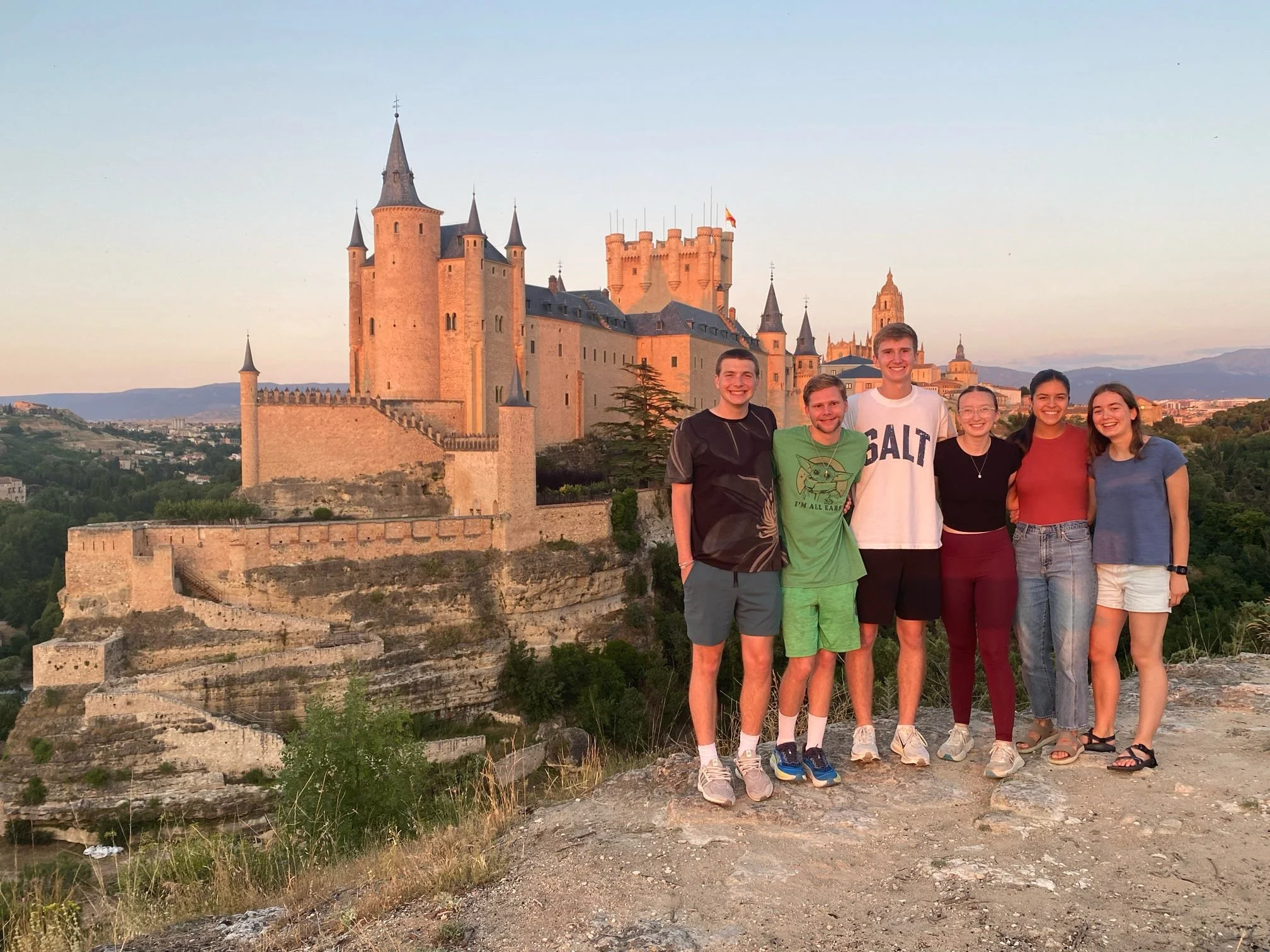 A group of seven young adults standing together in front of a large, medieval-style castle at sunset. The castle is built on a hill with a scenic background of rolling hills and distant mountains.