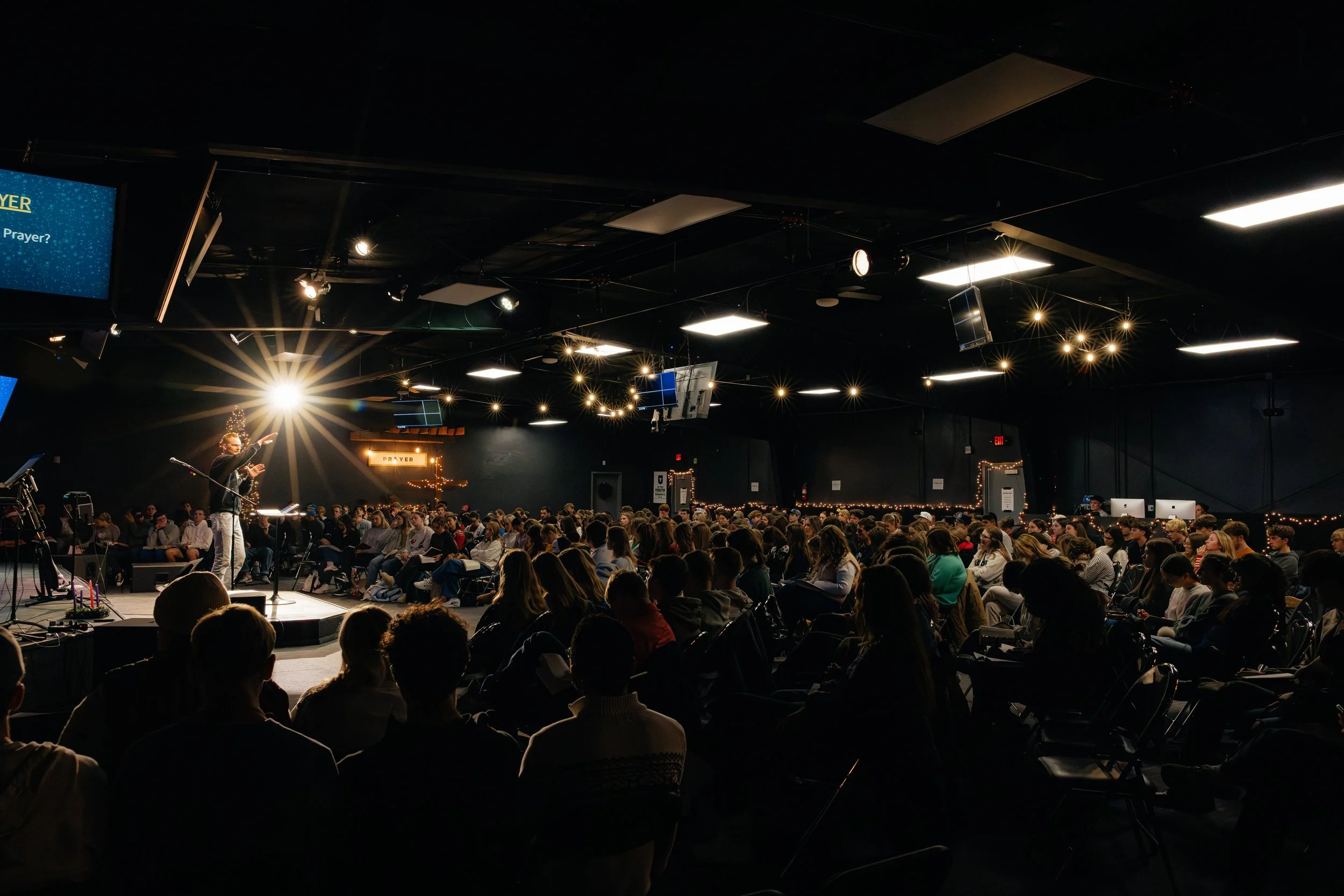A large indoor audience attending a prayer event with a stage, a speaker, and decorative lighting.