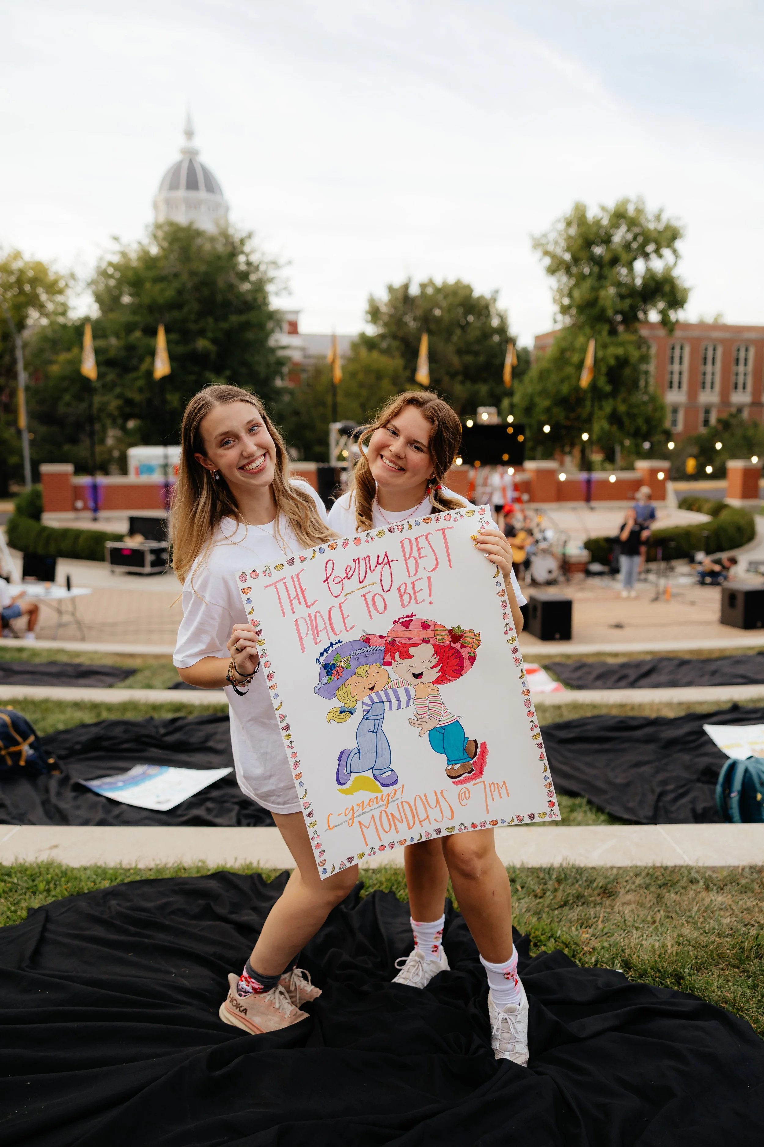 Two young women standing outdoors, holding a colorful sign that indicates a group event on Mondays at 7 PM; they are smiling and look happy, with a stage and trees in the background.