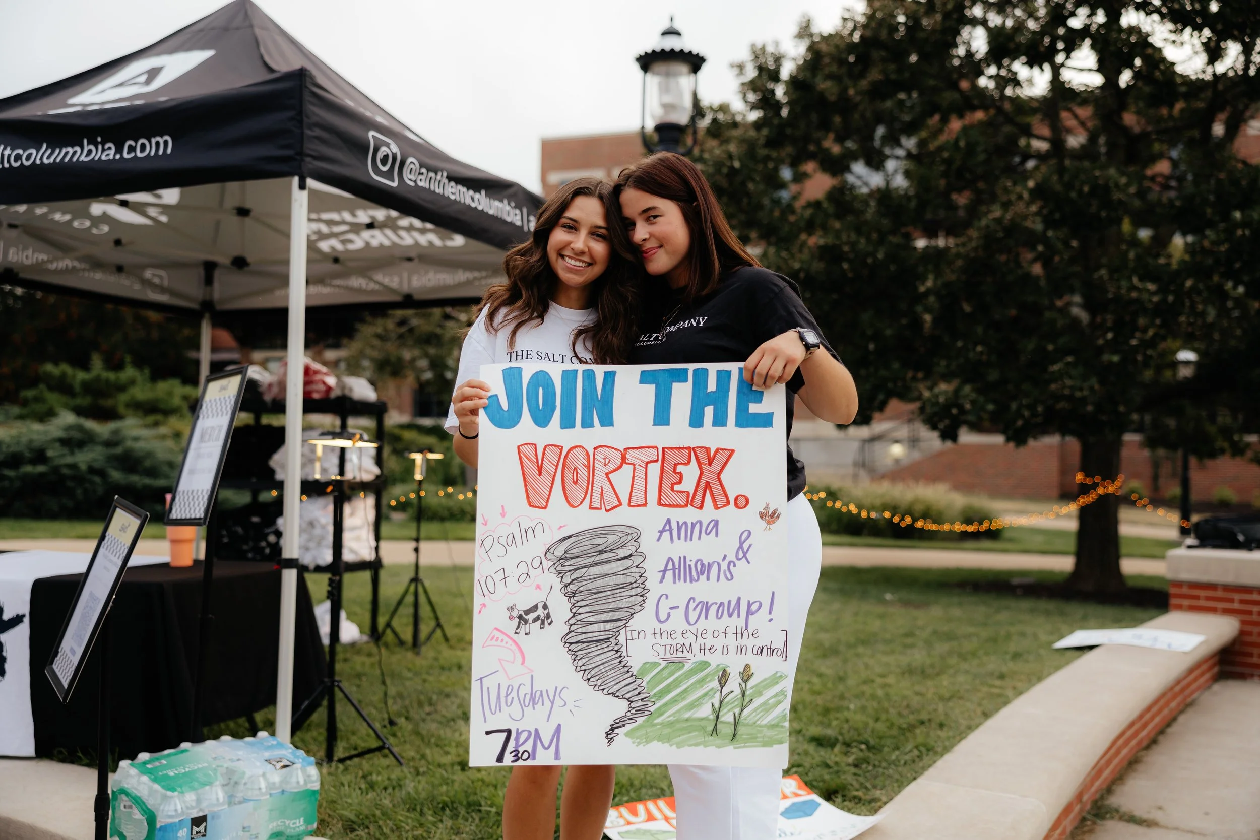Two young women smiling and holding a colorful sign that says "Join the Vortex" at an outdoor event, with a black canopy tent behind them and trees in the background.