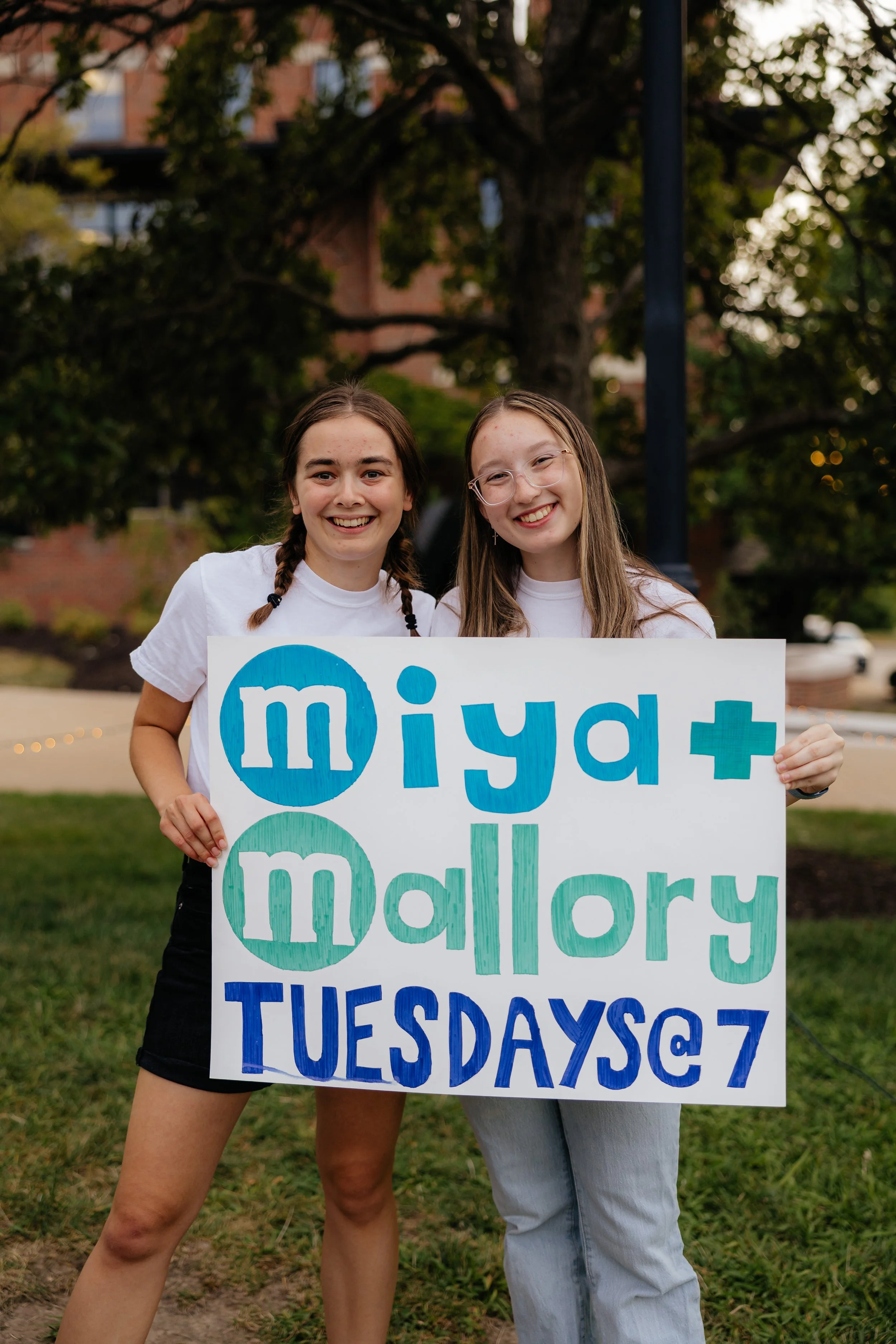 Two young women smiling and holding a large sign with colorful text that reads 'miyah mallory TUESDAYS @7', standing outdoors on grass with trees and a building in the background.