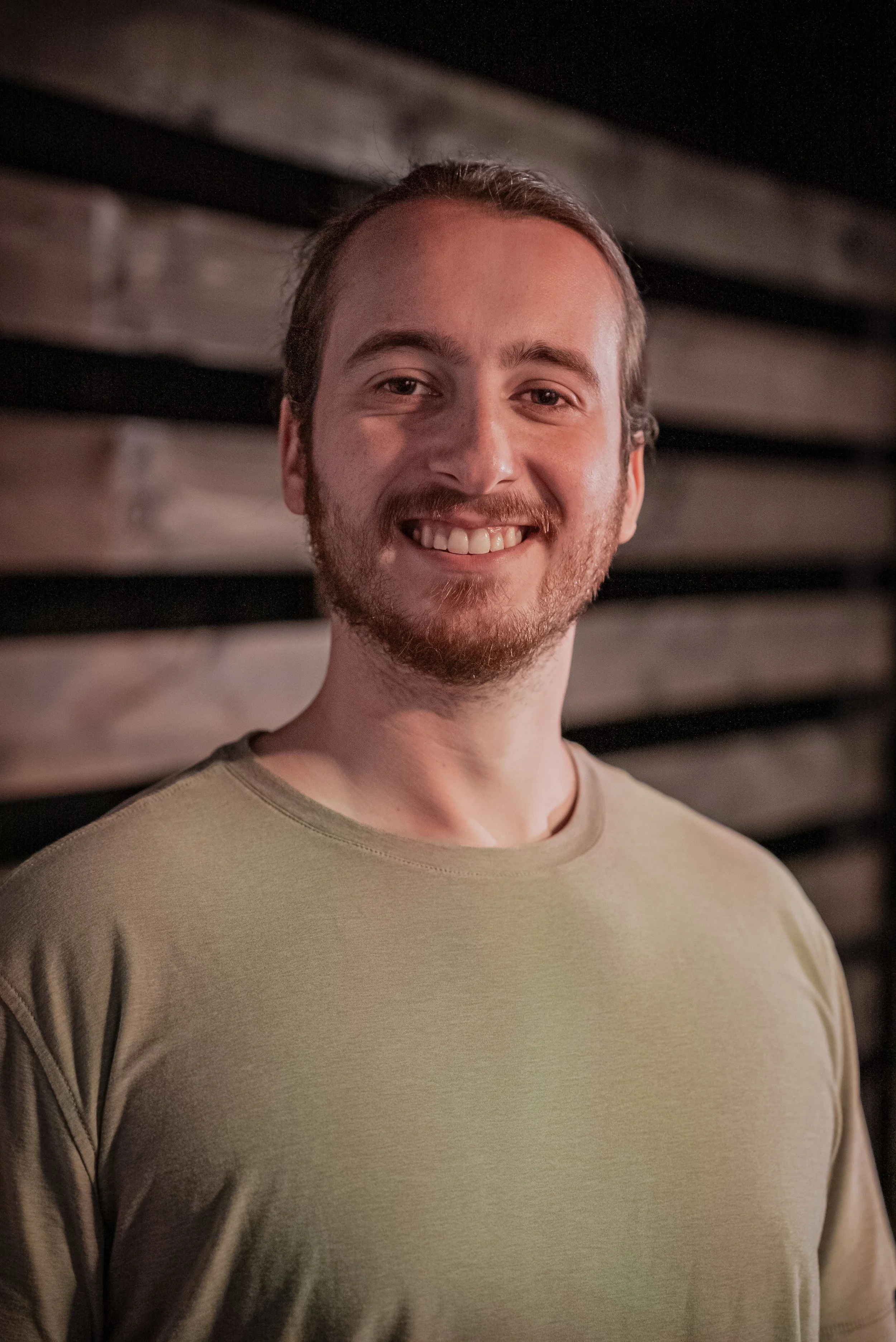 A smiling man with light brown hair and a beard, wearing a beige t-shirt, standing in front of a wooden wall.