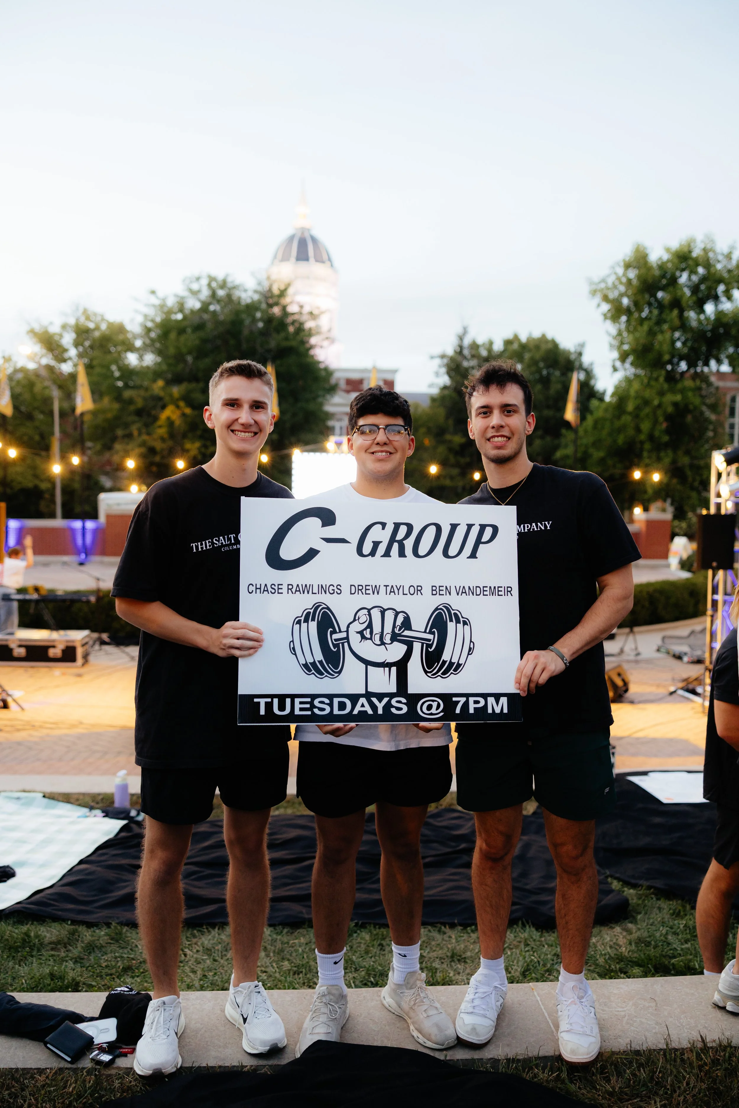 Three young men standing outdoors holding a sign promoting C-Group.