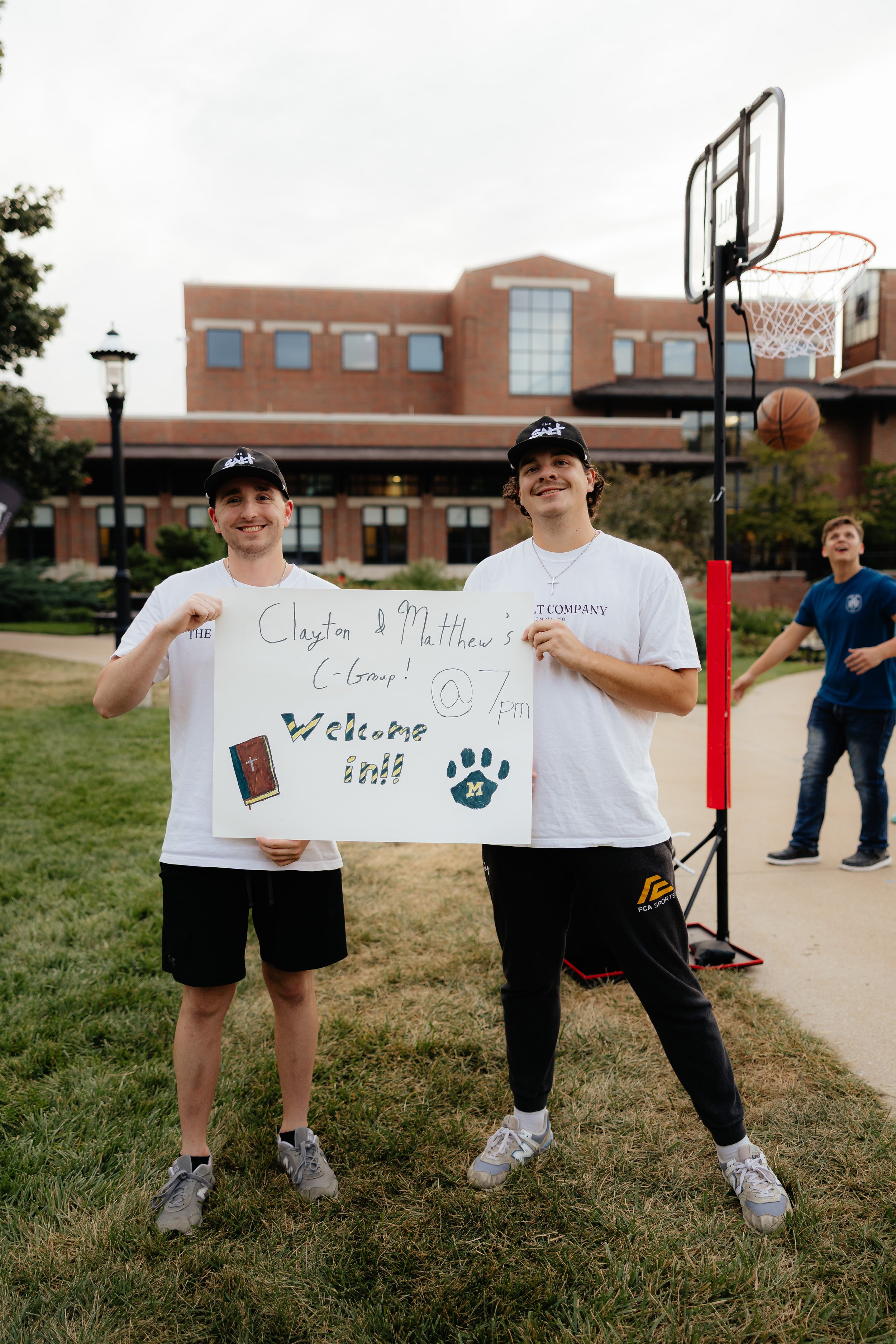 Two young men standing outdoors on a grassy area, holding a sign inviting others to join their group at 7 pm, with a basketball hoop and another person shooting hoops in the background.