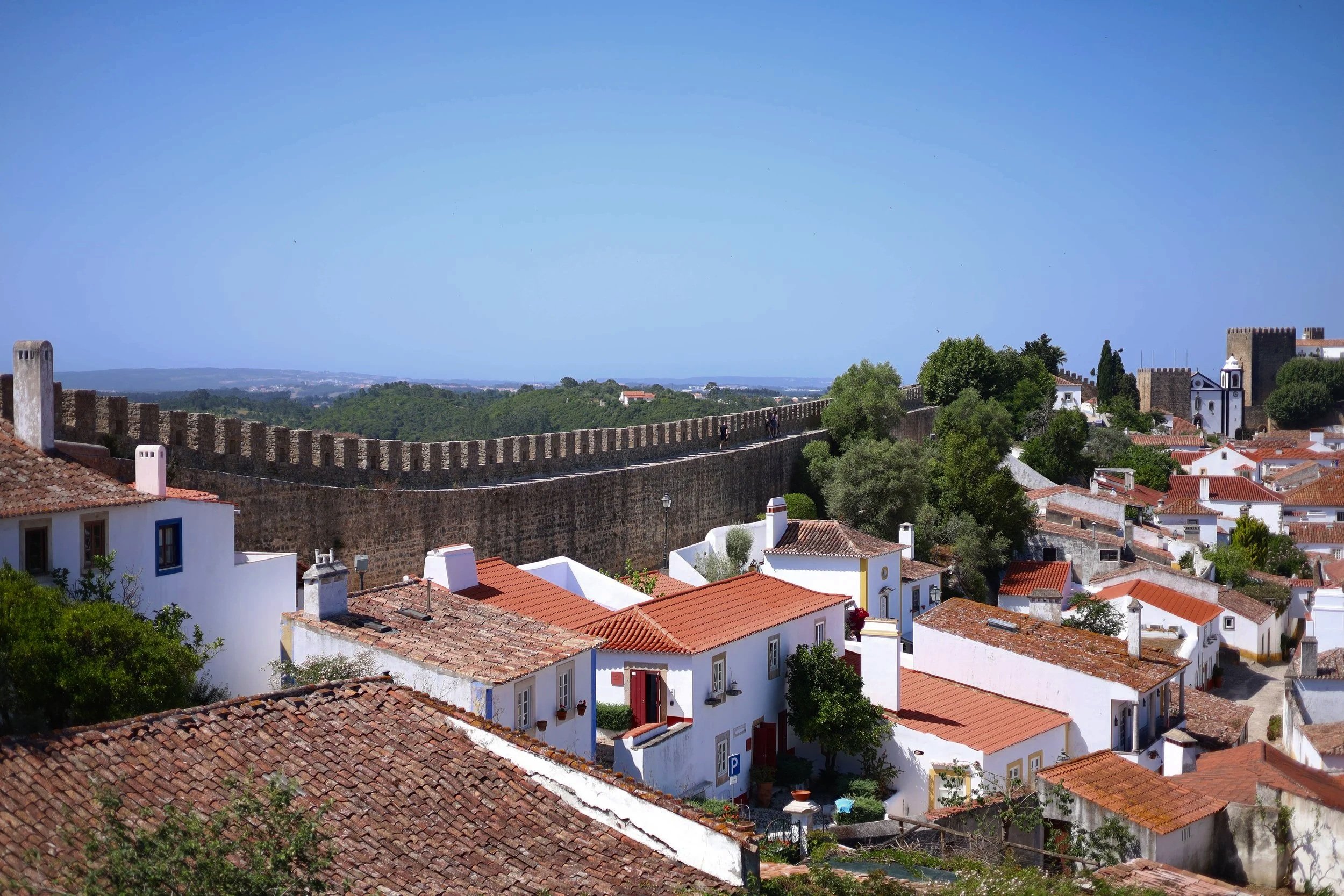 Scenic view of white houses with red-tiled roofs, a stone city wall, and lush green trees under a clear blue sky in a historic town.