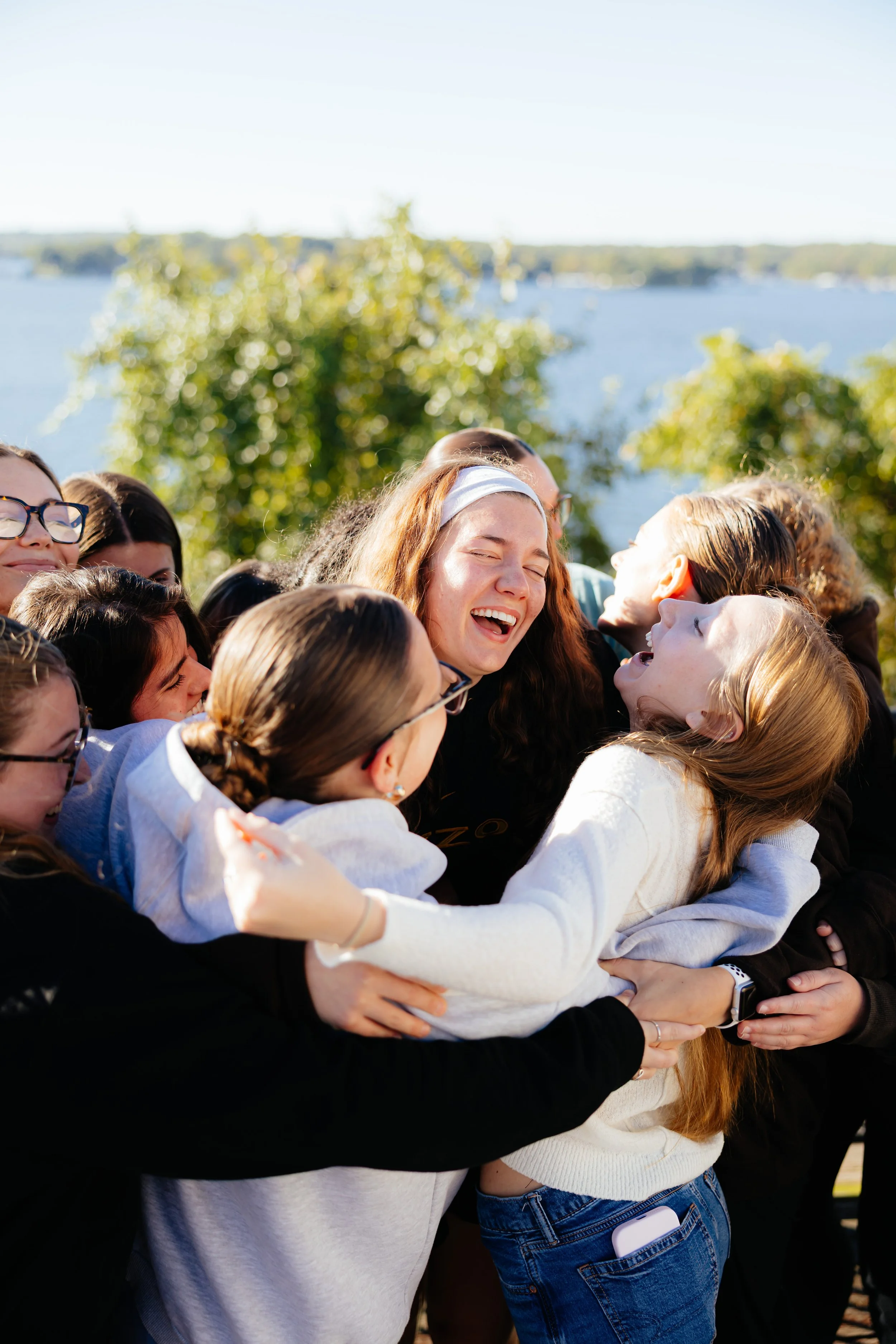 Group of smiling young women hugging outdoors near a lake with trees and boats in the background on a sunny day.