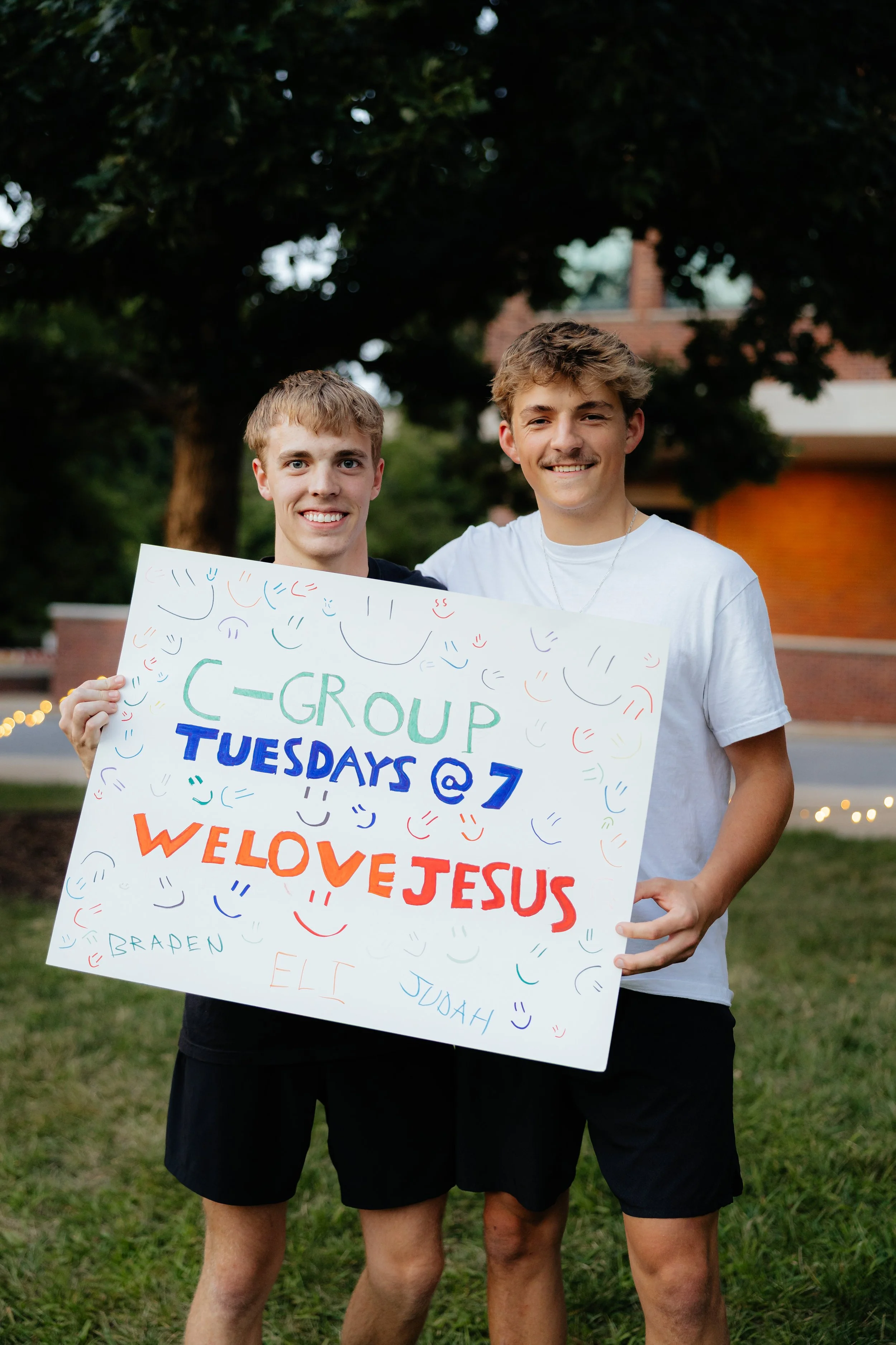Two young men standing outdoors at sunset, smiling and holding a colorful sign with text about a church group meeting. The background features trees, a brick building, and string lights.
