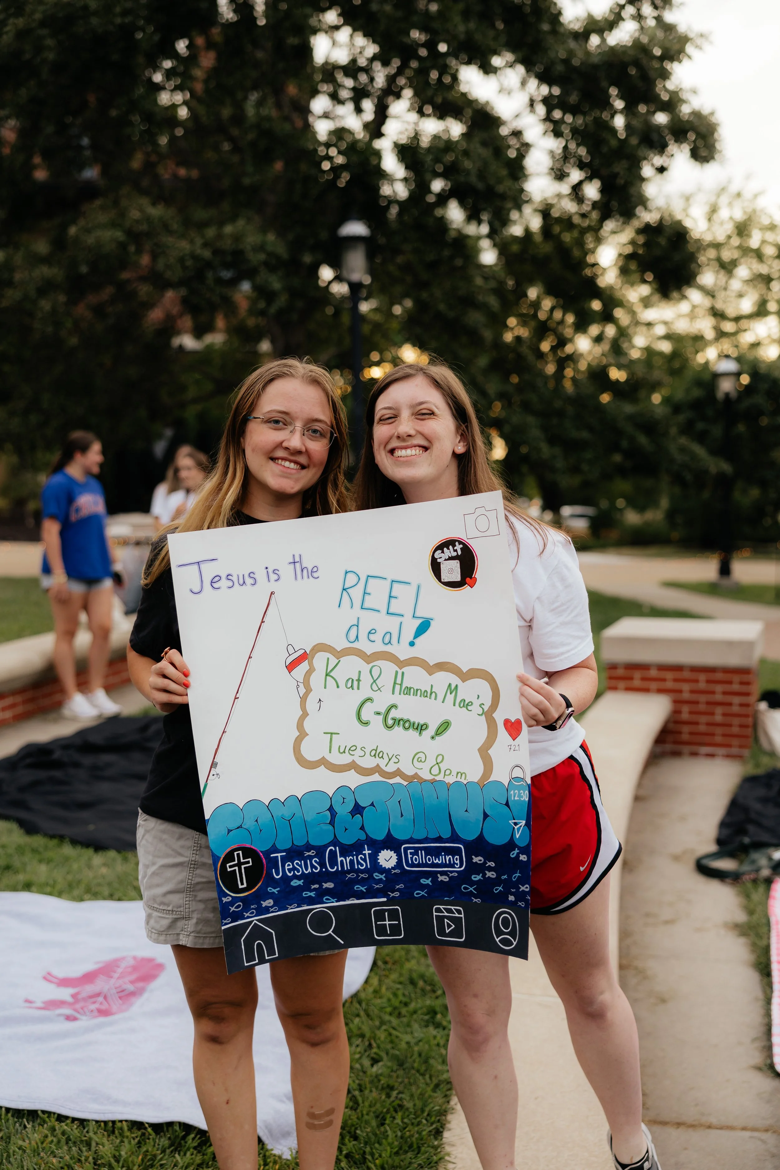 Two young women smiling and holding a colorful poster at an outdoor gathering, with other people in the background and trees in a park setting.