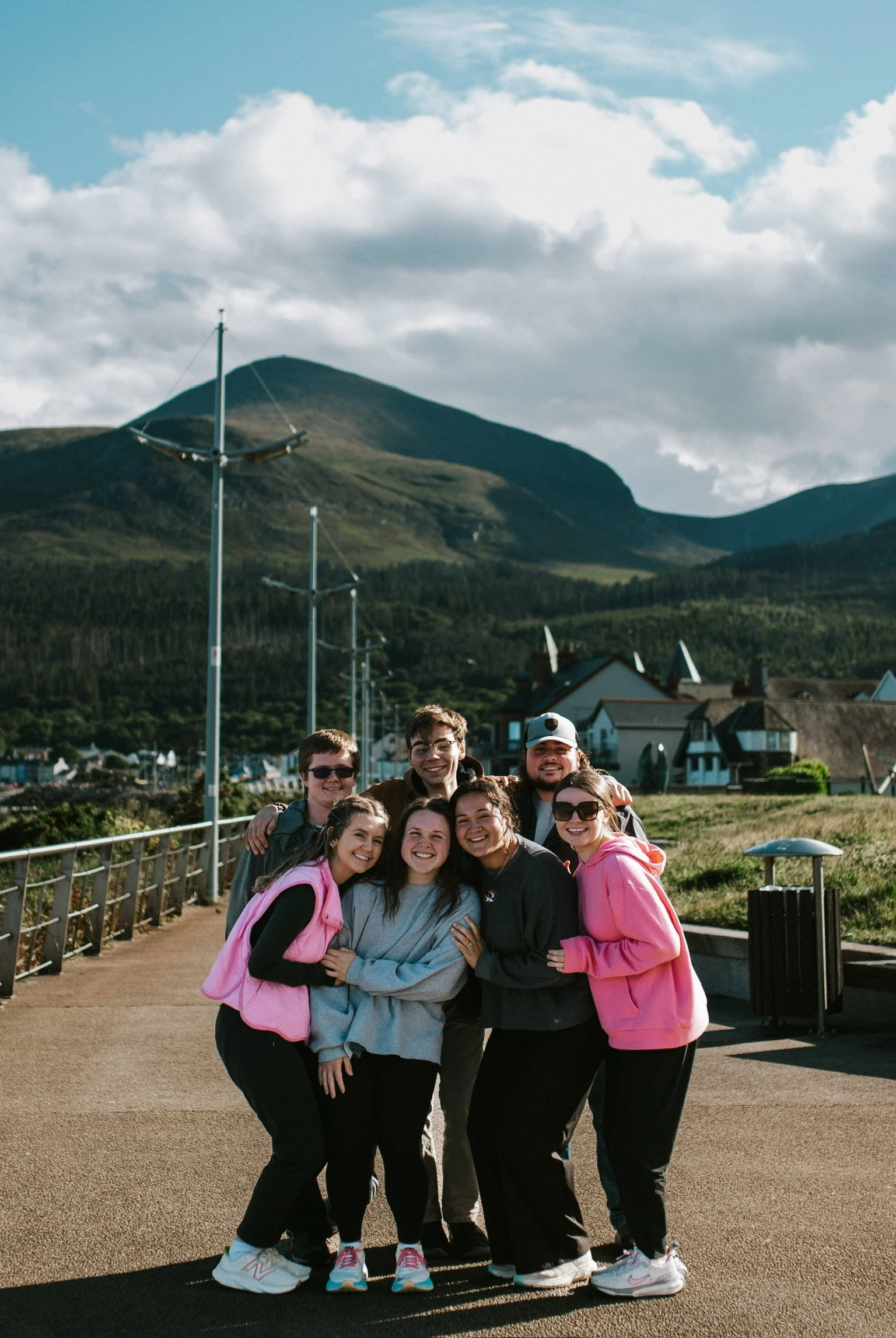 A group of six friends smiling and hugging outdoors with mountains, houses, and blue sky in the background.