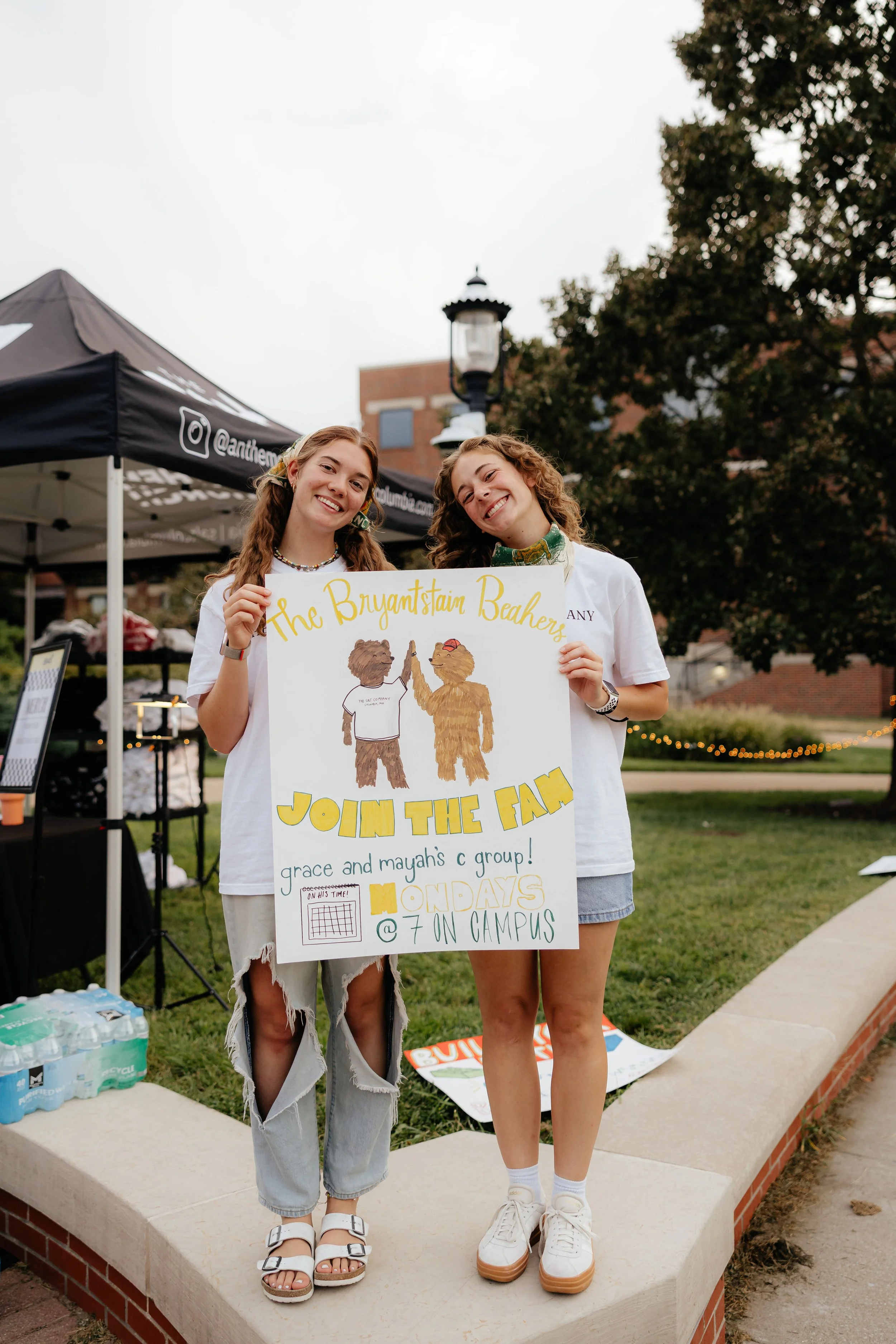Two young women standing outdoors holding a colorful poster promoting a campus event, smiling at the camera.