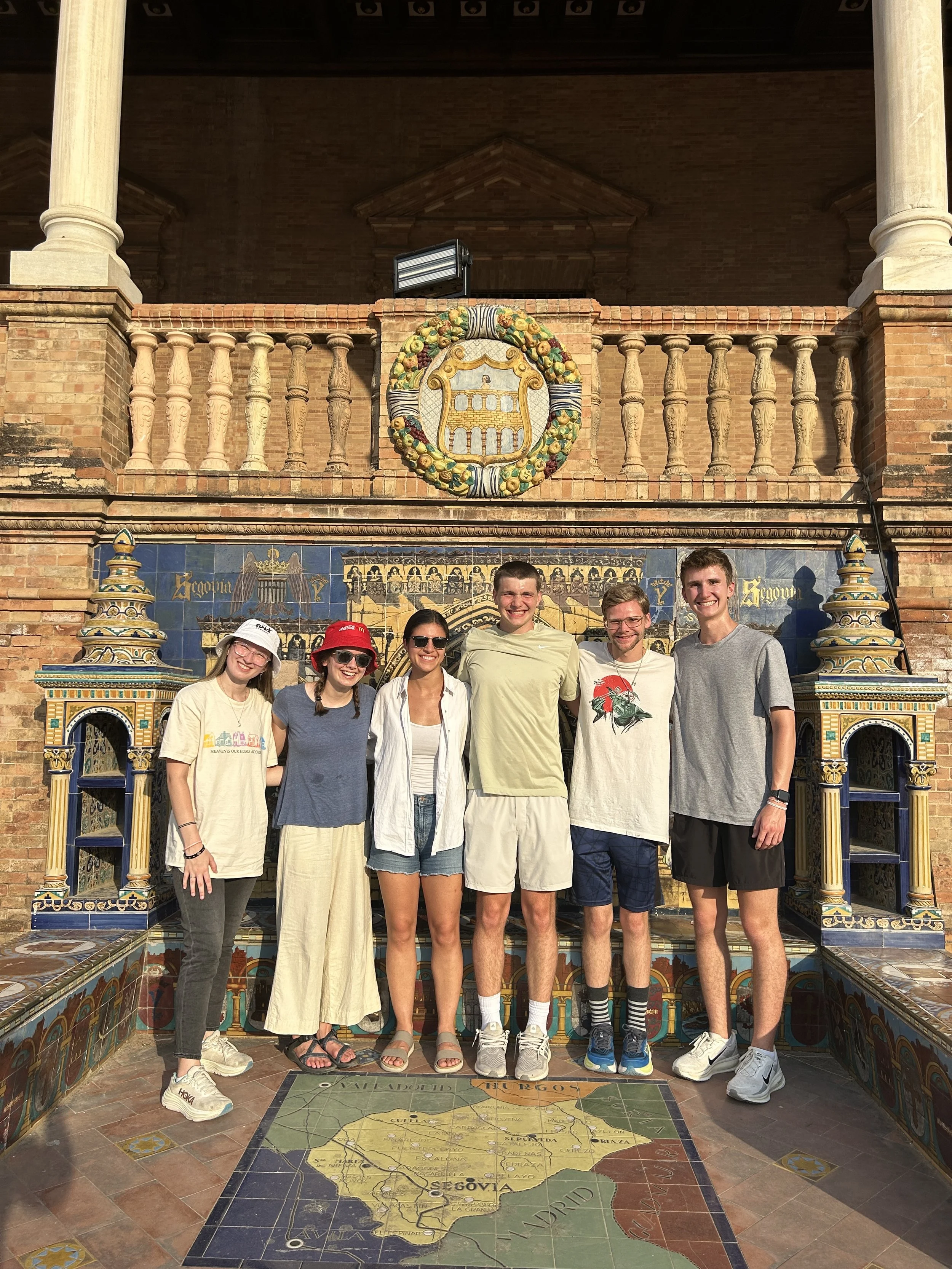 Six young people standing on a colorful tiled floor with a map of Spain, in front of decorative tile and brickwork, smiling at the camera.