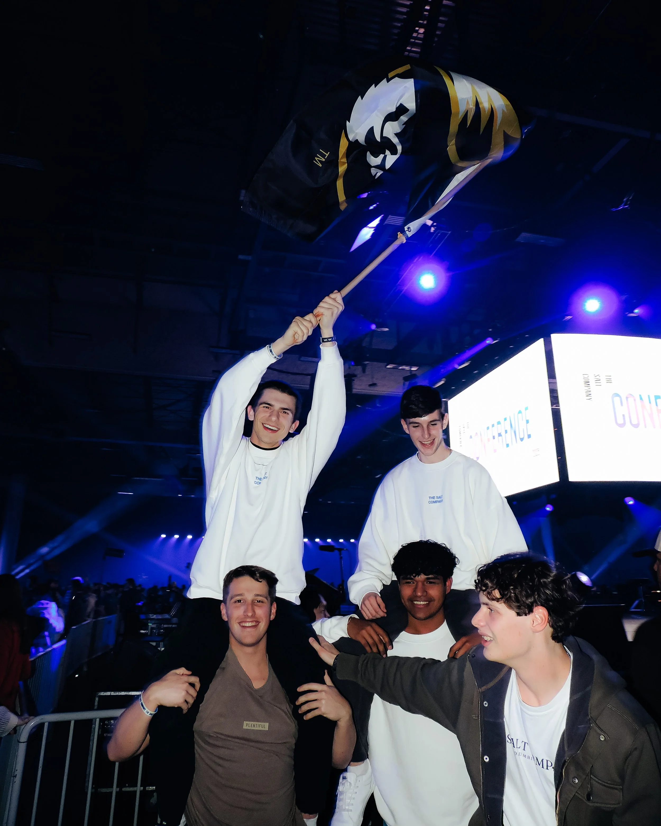 Group of young men celebrating at an event, one holding a flag with a lion logo, standing on others' shoulders, in an indoor venue with stage lights and screens in the background.