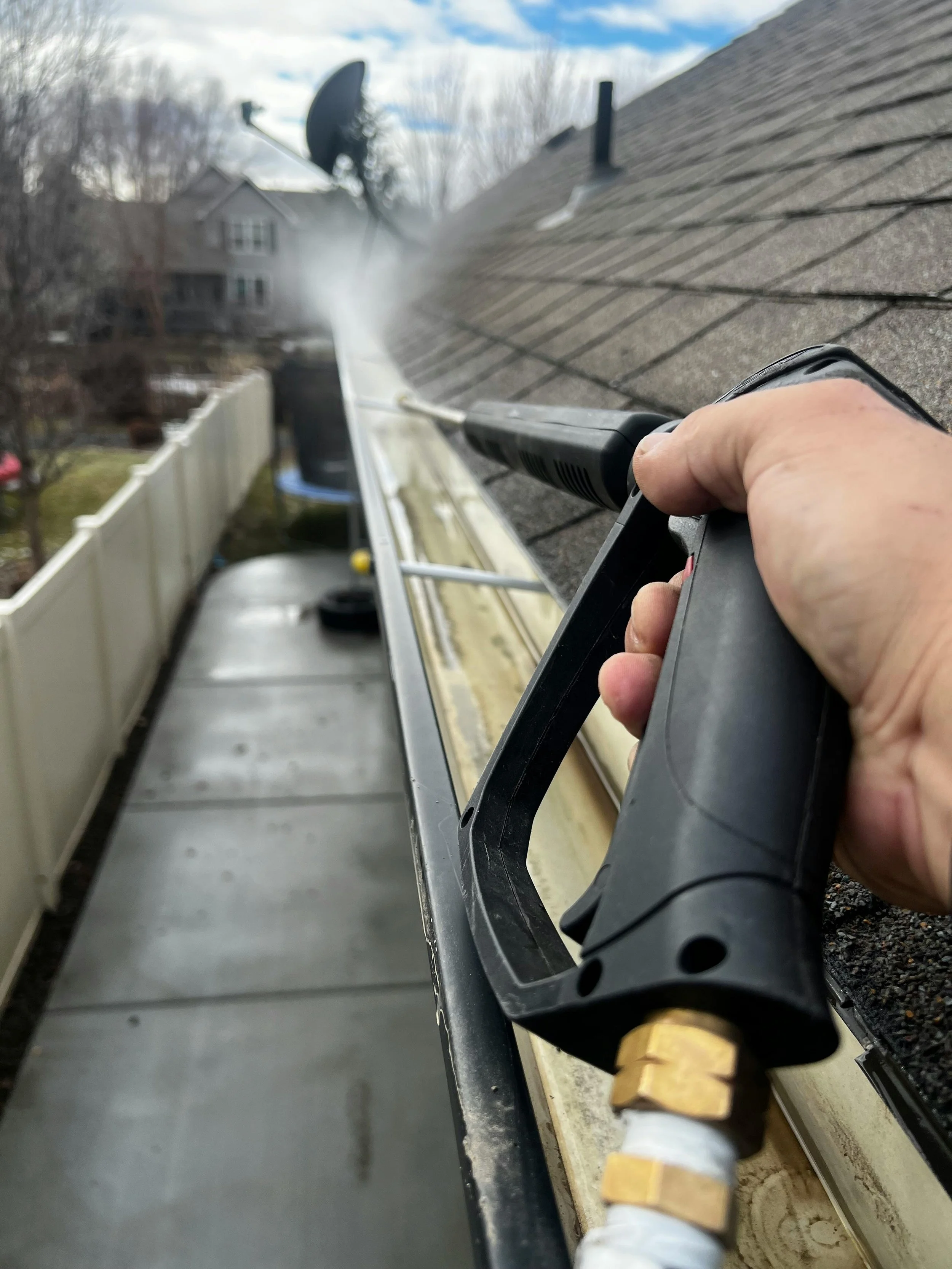 A person using a power washer to clean the roof of a house with a spray of water, with trees and a house in the background.