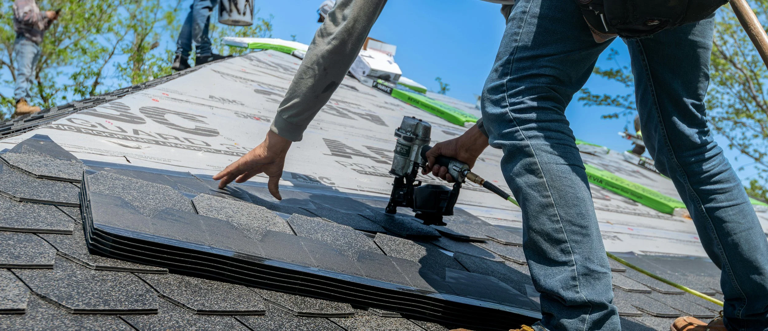 Worker installing or repairing shingles on a roof with roofing tools and materials, under a clear blue sky.