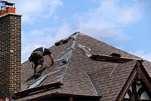 A worker on a roof repairing or installing shingles on a house in Montreal with a brick chimney, under a blue sky.